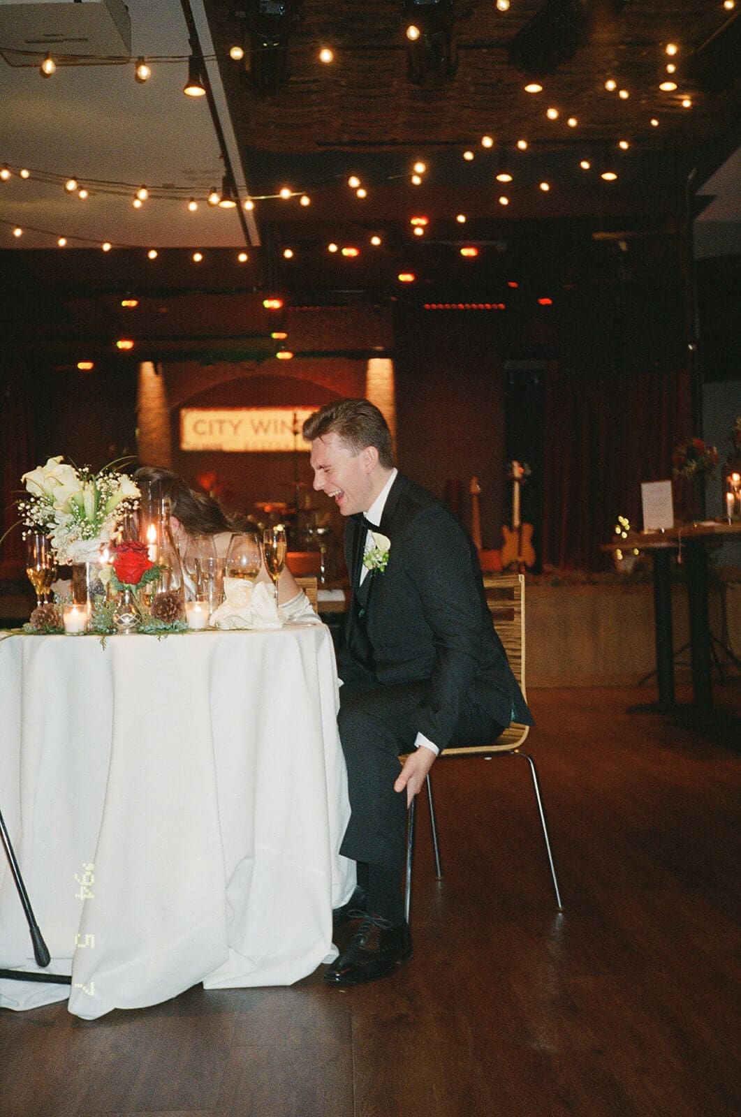 Candid wedding photos of the groom laughing at the sweetheart table during a reception speech.