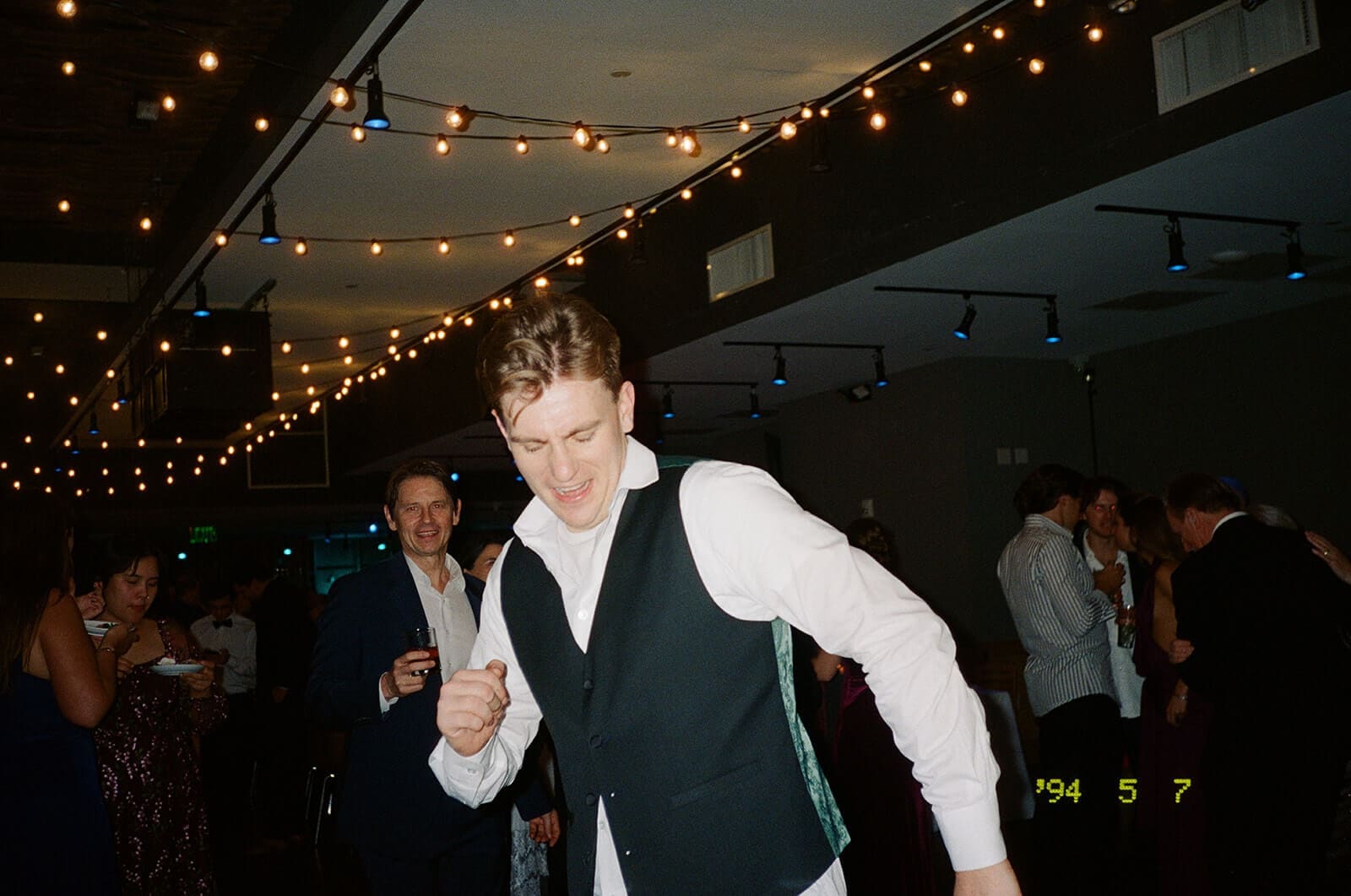 Candid wedding photos of a guest in a vest dancing energetically beneath the reception string lights.