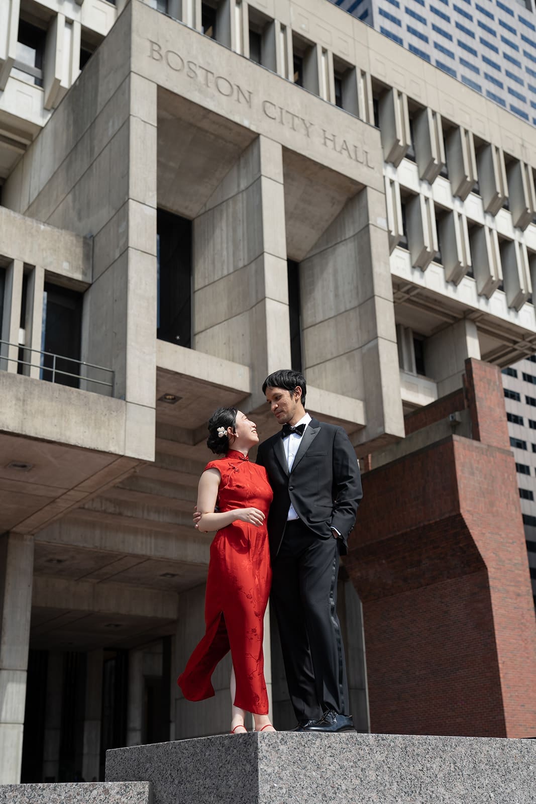 Couple in a red dress and black tuxedo poses outside Boston City Hall.