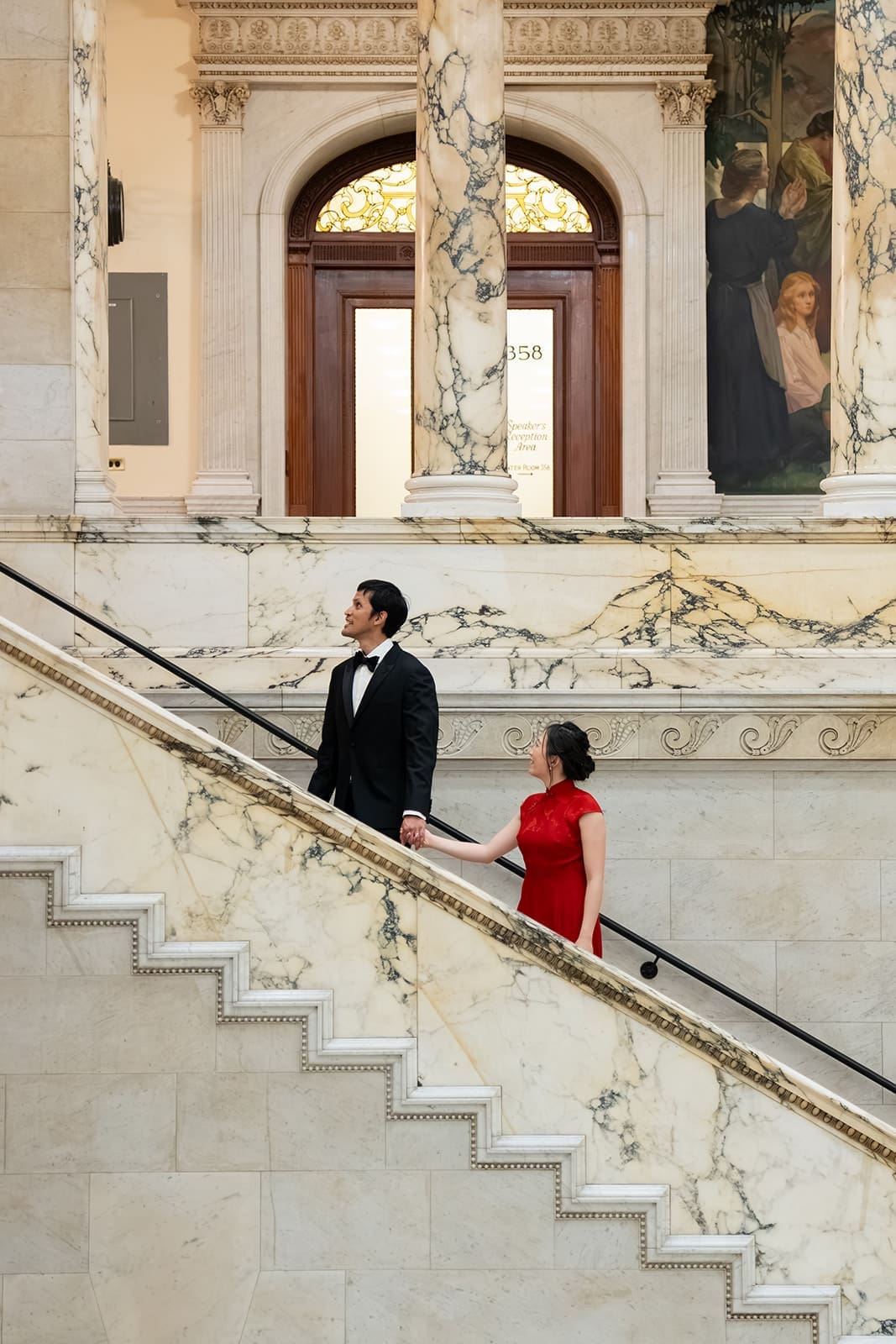 Newlyweds walk hand in hand along the marble staircase inside Boston City Hall.