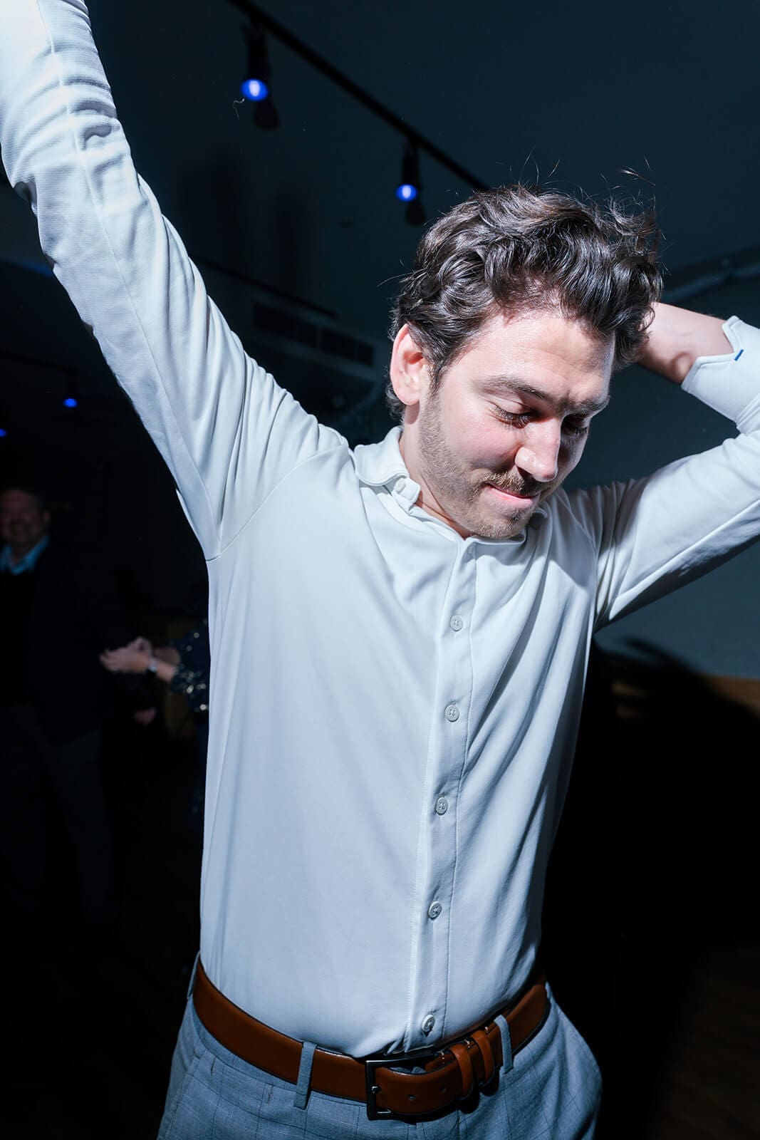 Candid wedding photos of a wedding guest dancing with his arms raised beneath colorful reception lighting.