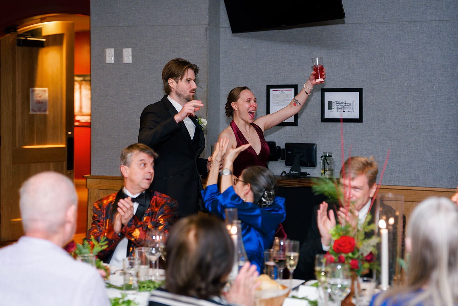 Candid wedding photos of two wedding party members raising their drinks while guests applaud.