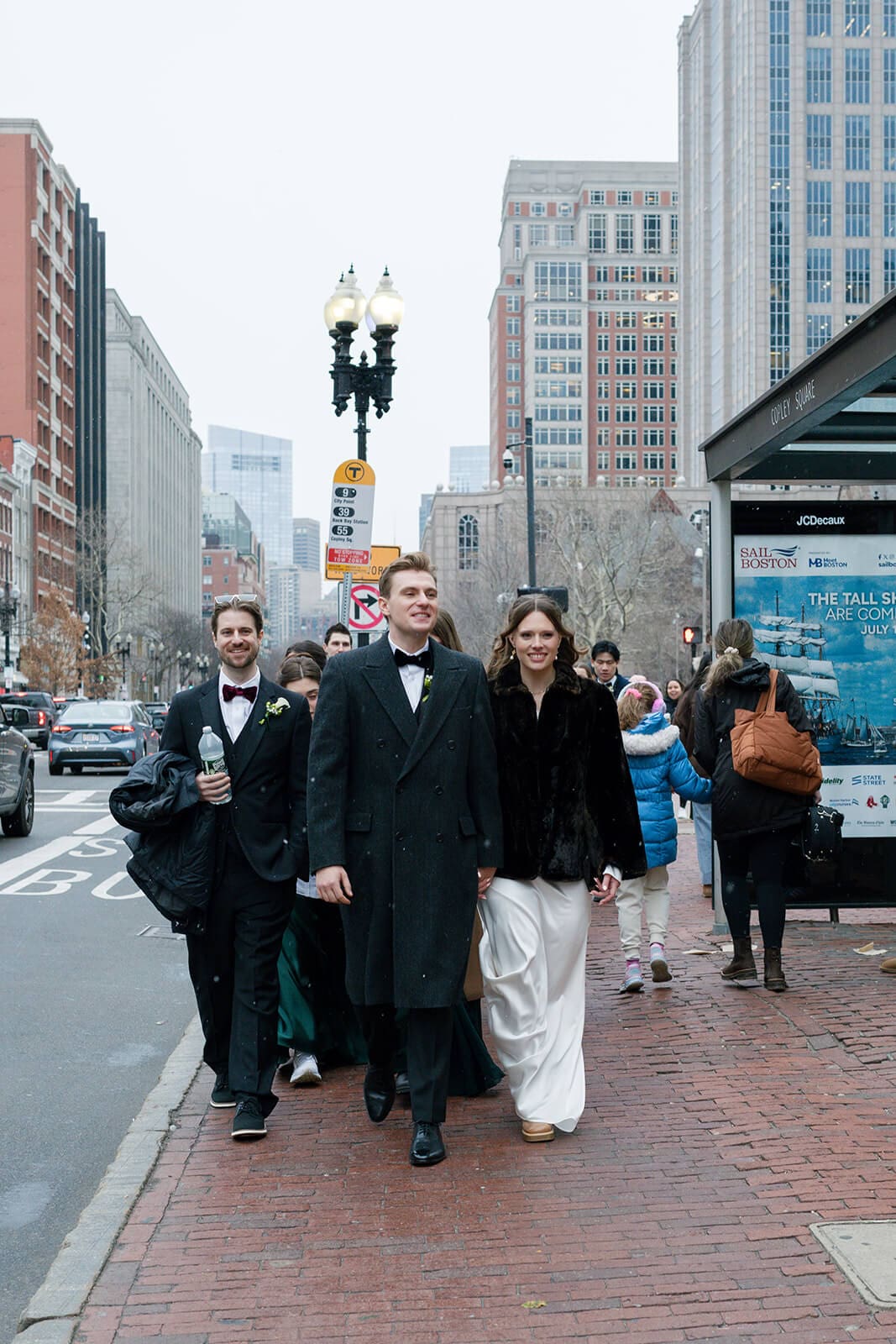 Bride and groom walking with their wedding party along a brick sidewalk near Copley station in Boston.