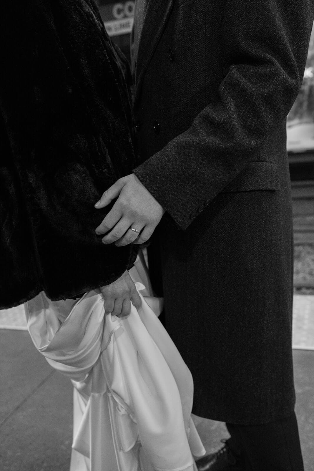Black and white close-up of newlyweds holding hands, showing the bride’s wedding ring and gathered dress.