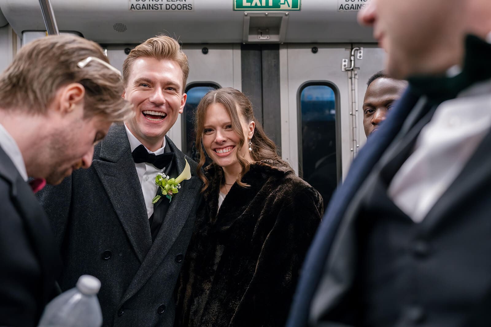 Bride and groom laughing with friends aboard a Boston MBTA train during their wedding day celebration.
