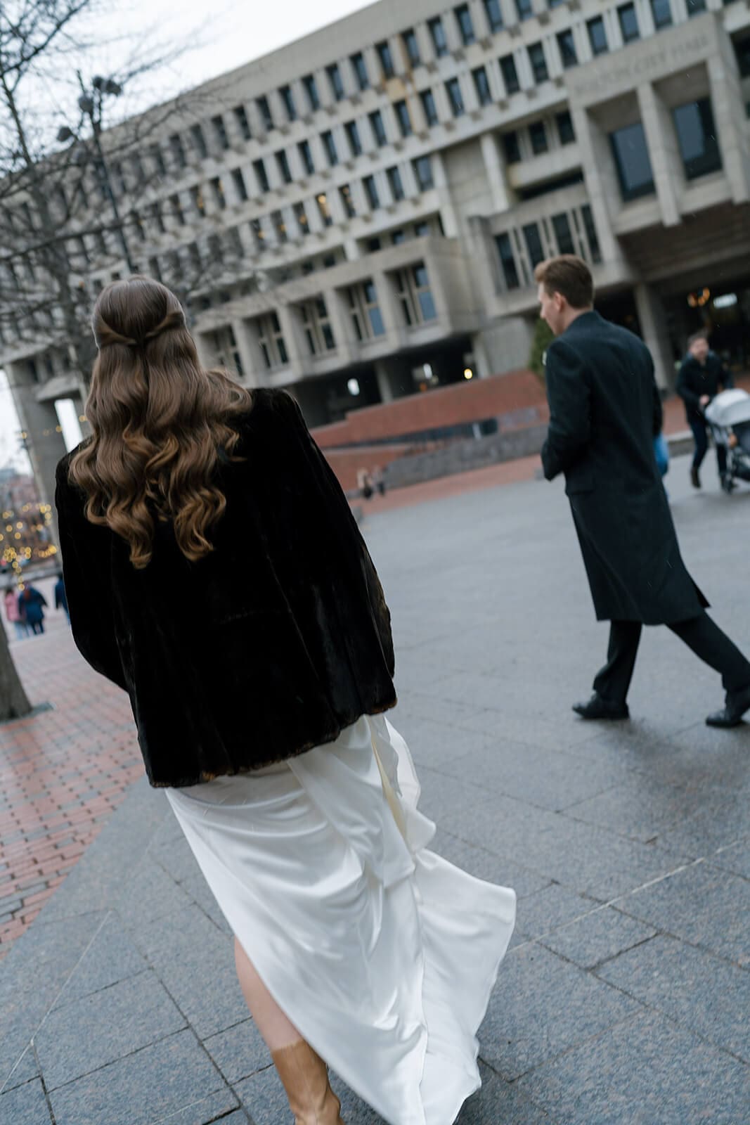 Bride walking across a Boston plaza with her wedding dress gathered as city buildings rise behind her.