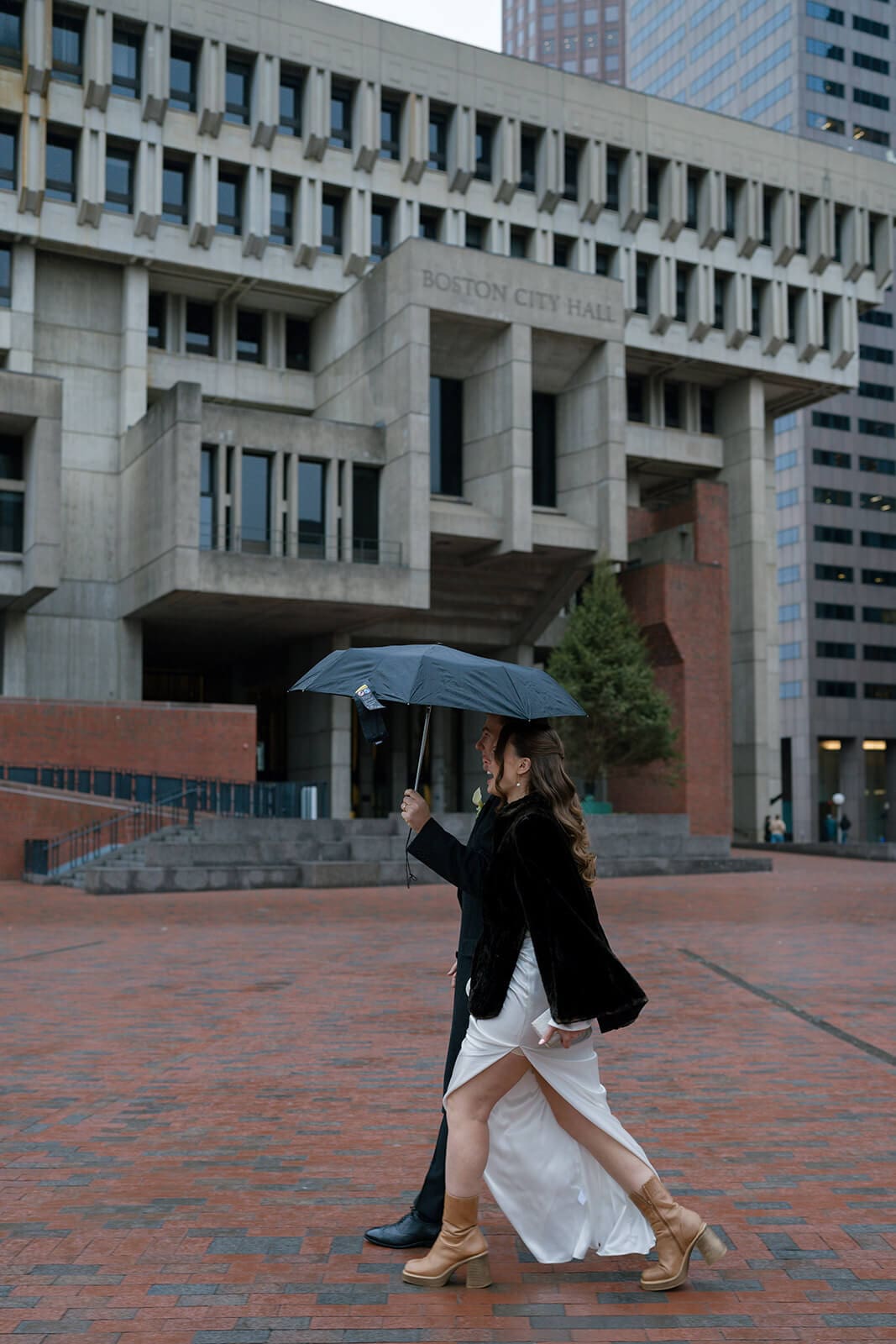 Bride walking through a downtown Boston plaza beneath an umbrella with her wedding dress gathered in one hand.
