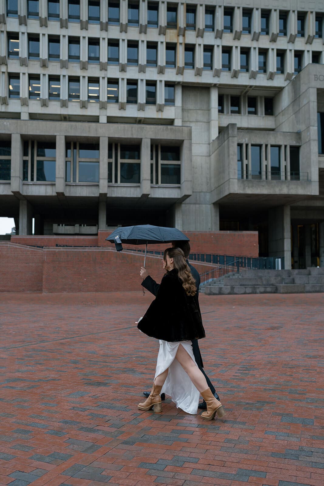 Bride walking across a downtown Boston plaza beneath an umbrella on her rainy wedding day.
