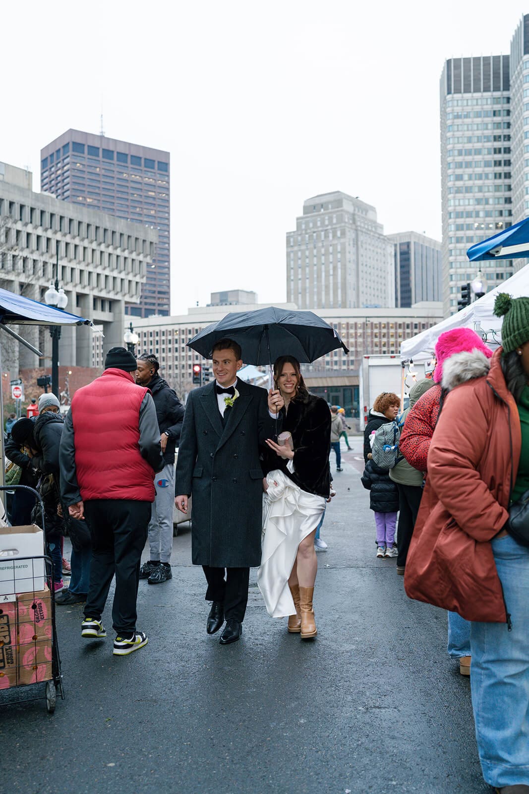 Bride and groom walking together beneath an umbrella through a crowded Boston outdoor market.