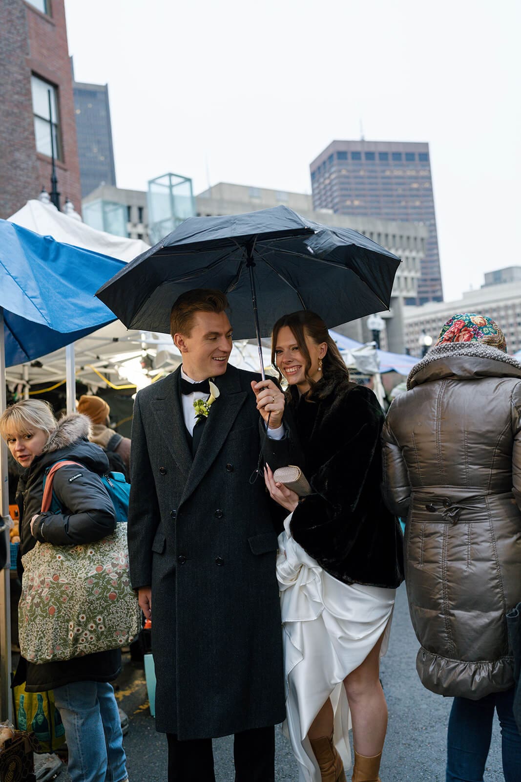 Newlyweds walking beneath an umbrella through a busy Boston market after their wedding ceremony.