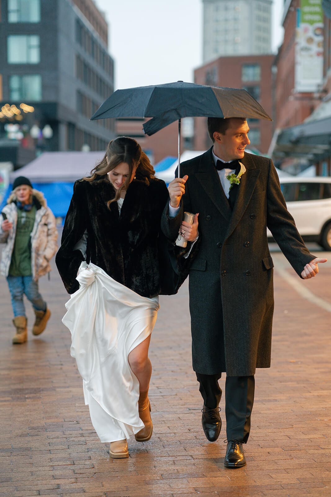 Newlyweds walking together beneath an umbrella along a rainy brick sidewalk in downtown Boston.