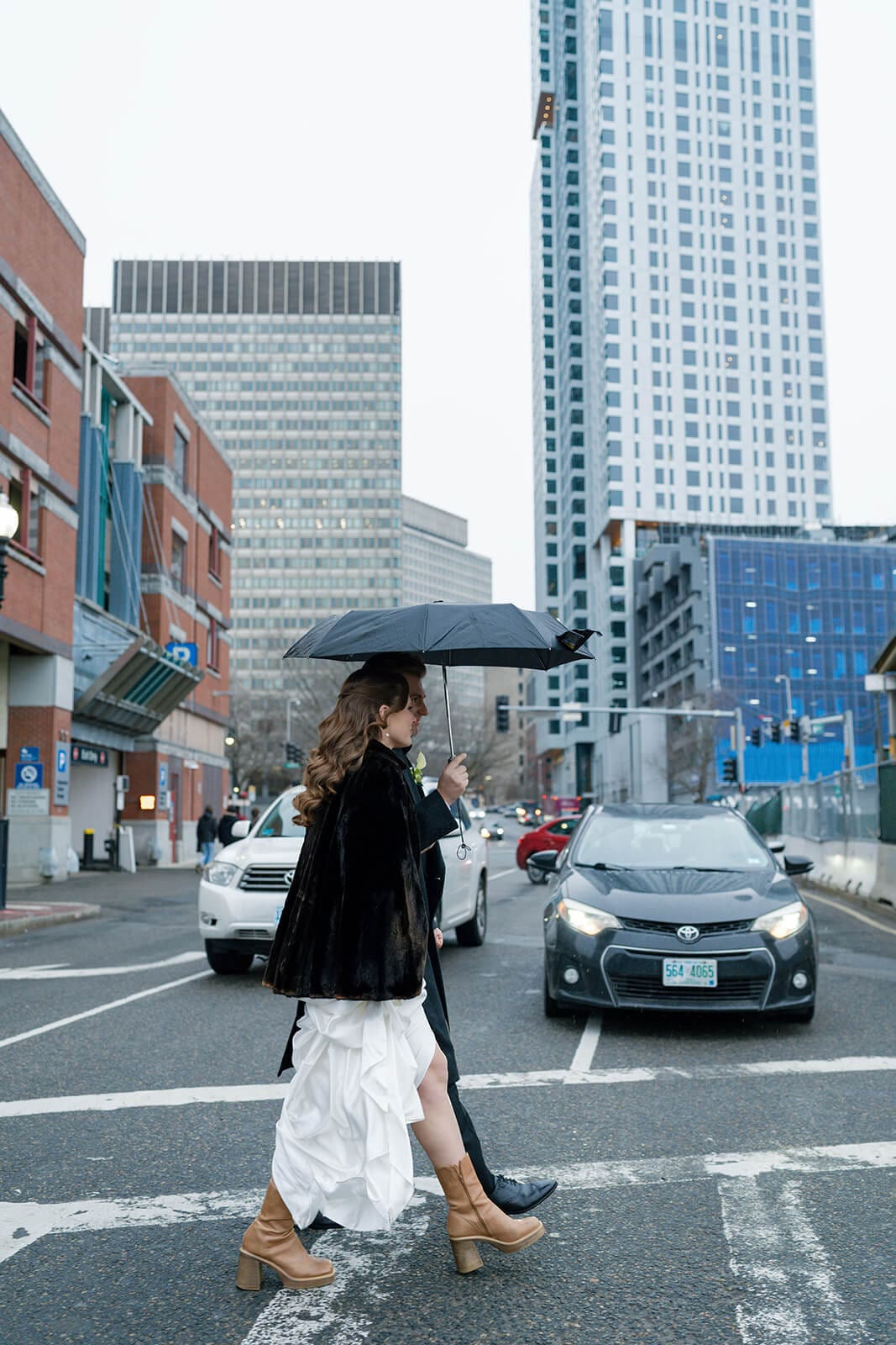 Bride walking through a Boston crosswalk beneath an umbrella with her wedding dress gathered in one hand.