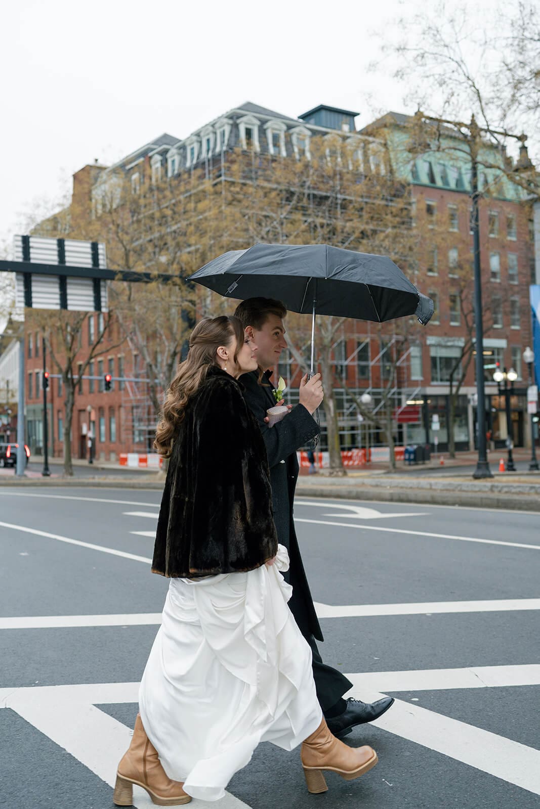 Bride and groom walking beneath an umbrella across a rainy Boston street on their wedding day.
