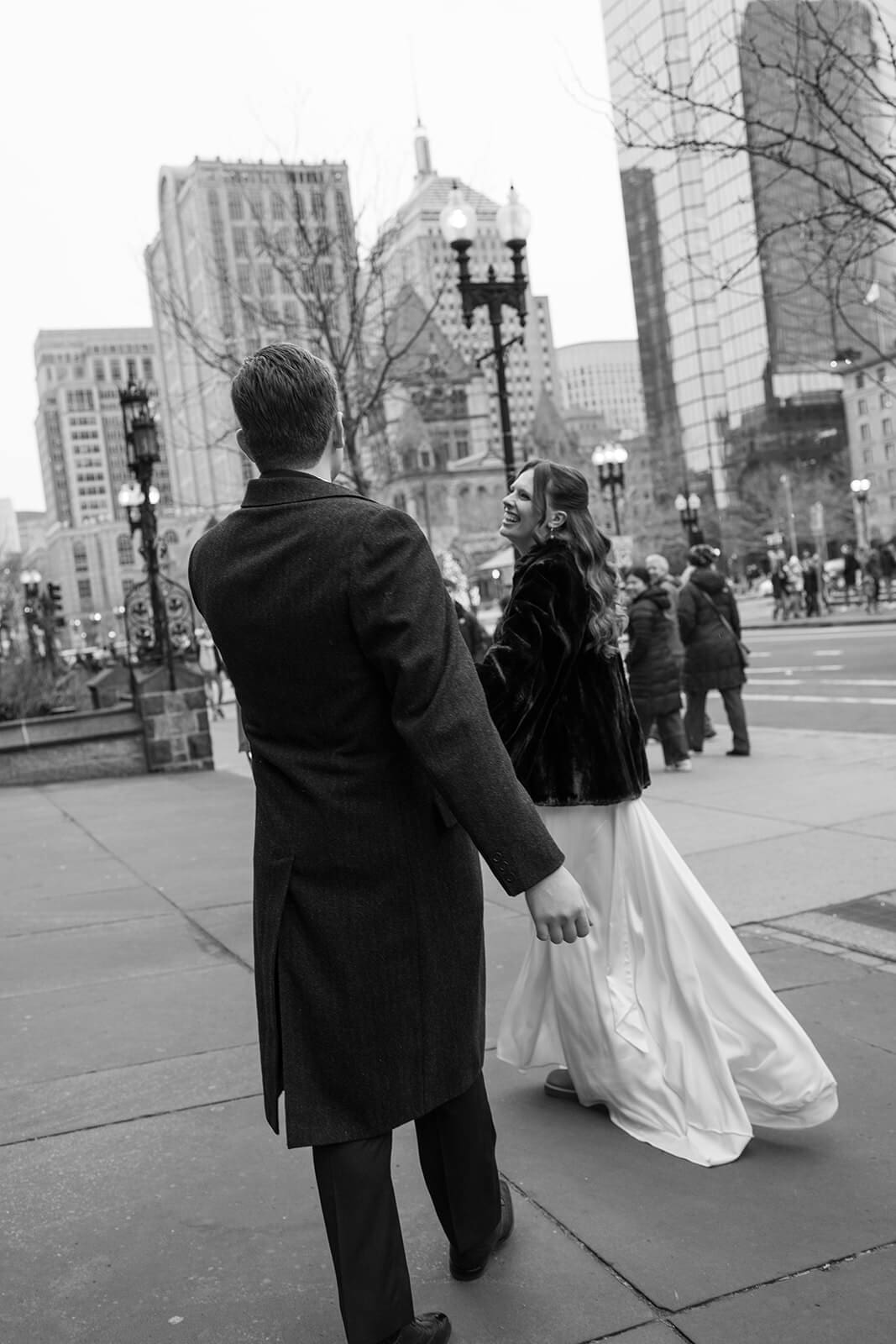 Black and white photo of a bride and groom walking through downtown Boston with the bride’s dress trailing behind her.