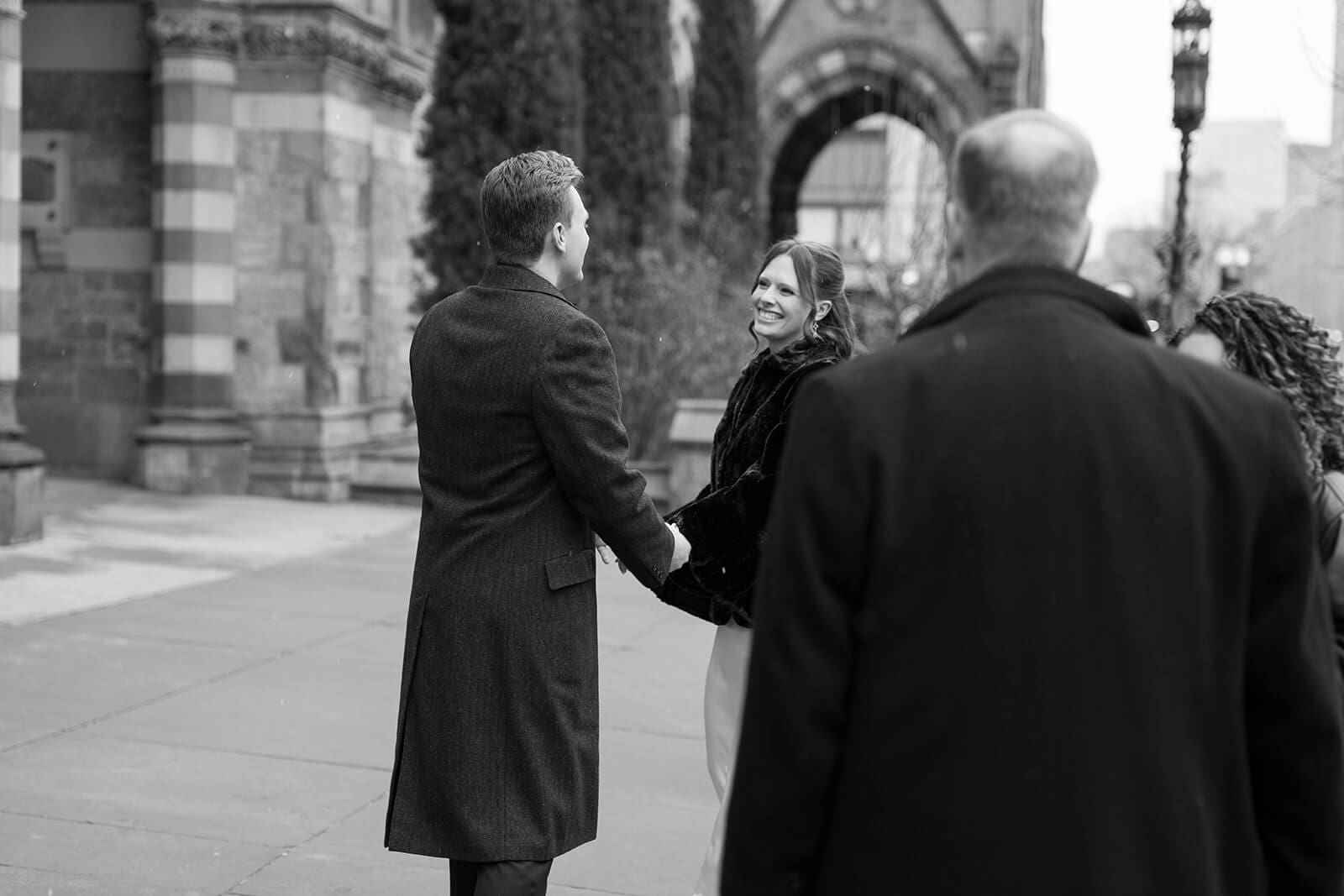 Black and white candid photograph of newlyweds holding hands and smiling outside a historic Boston church.