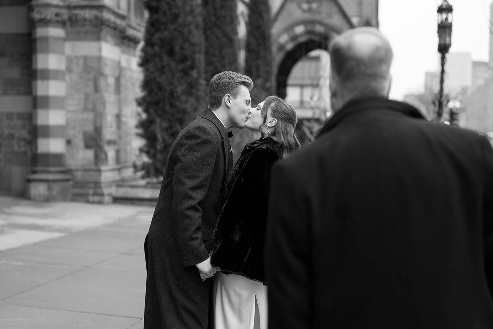 Black and white photograph of newlyweds kissing on a Boston sidewalk as a passerby crosses the foreground.