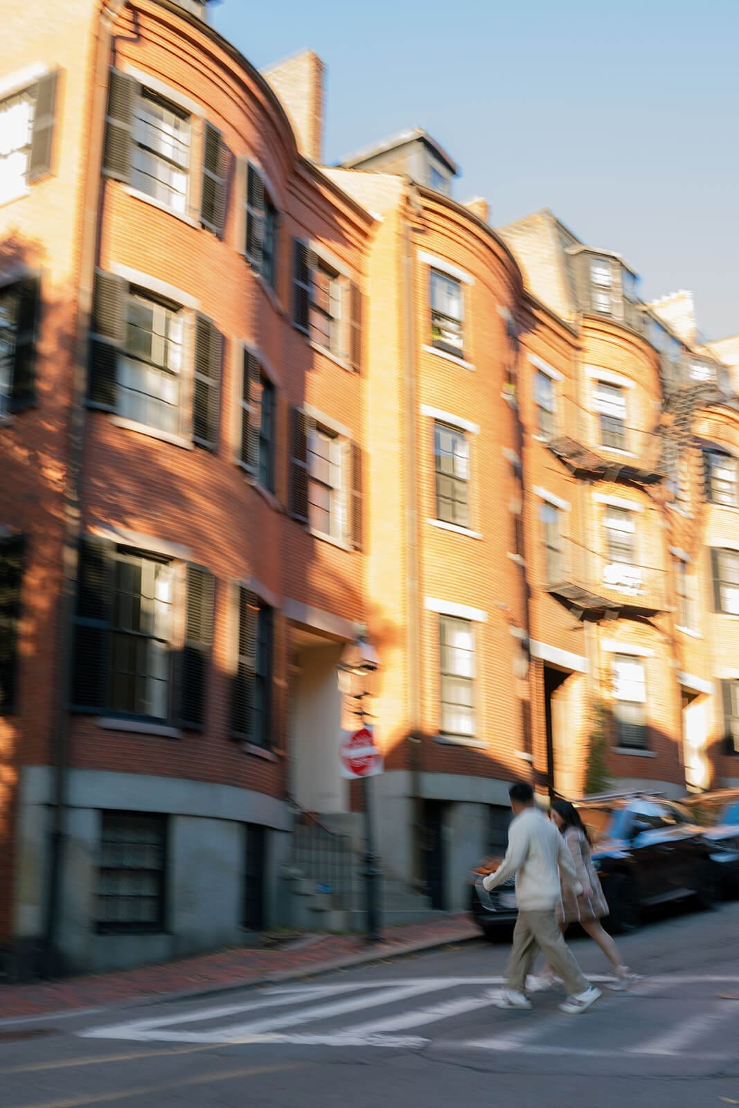 Motion-blurred photograph of a couple holding hands while crossing a street lined with historic Beacon Hill brick homes.