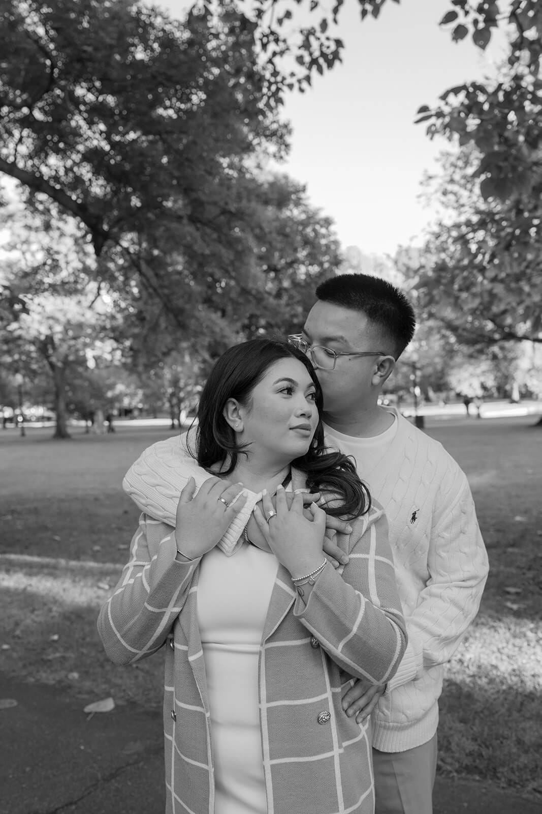 Black-and-white close portrait of a couple embracing, with the man standing behind his partner beneath the trees.