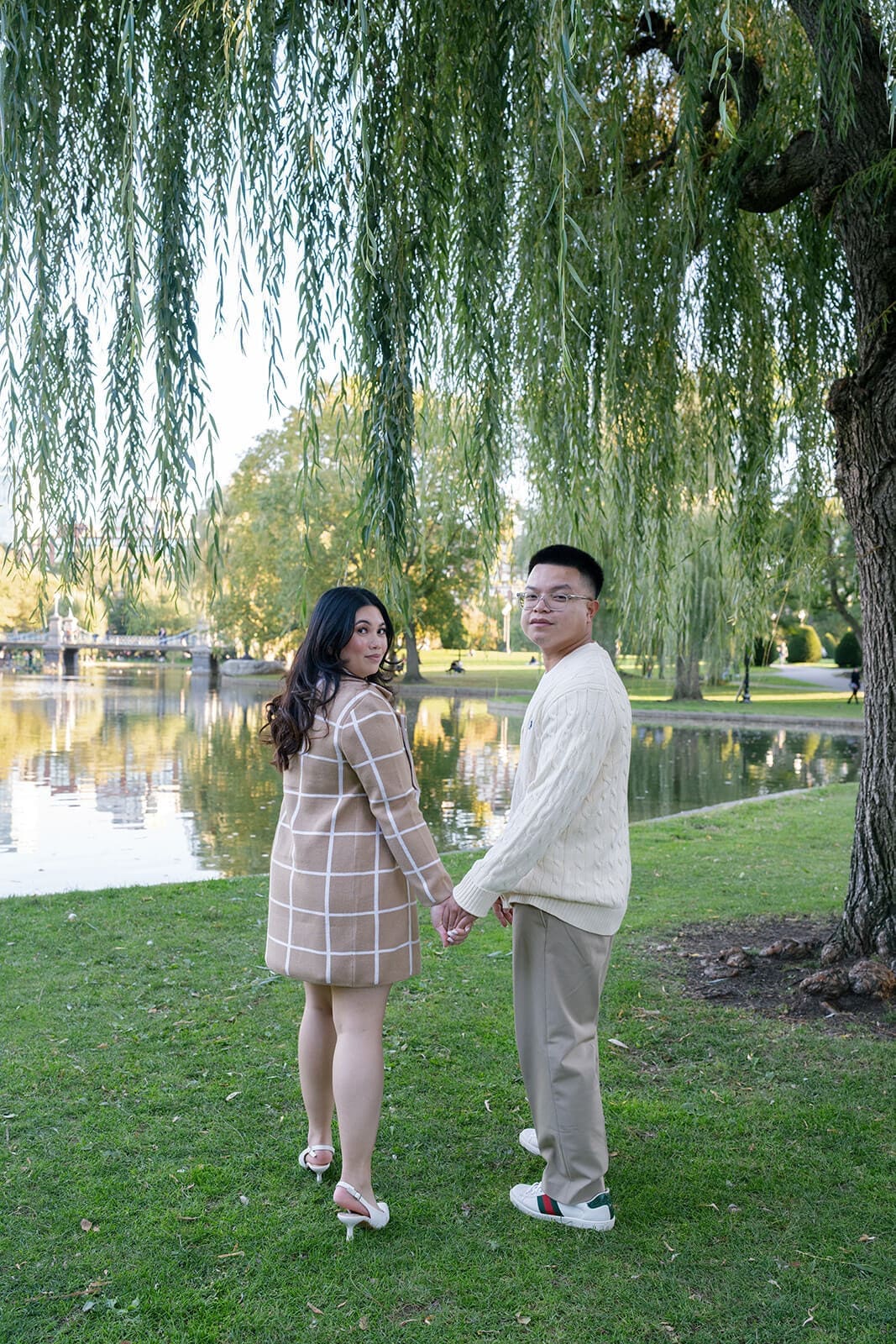Couple holding hands beside the pond in Boston Public Garden beneath the hanging branches of a willow tree.