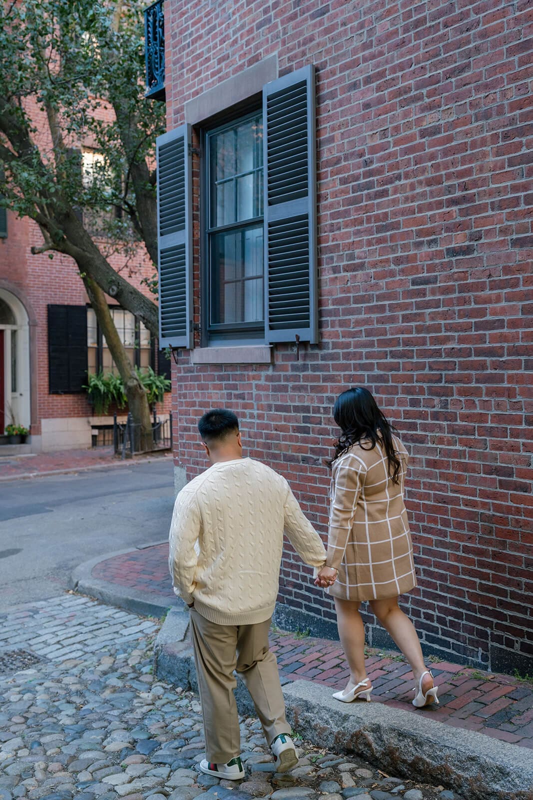 Couple holding hands while walking away along a cobblestone street surrounded by Beacon Hill brick buildings.