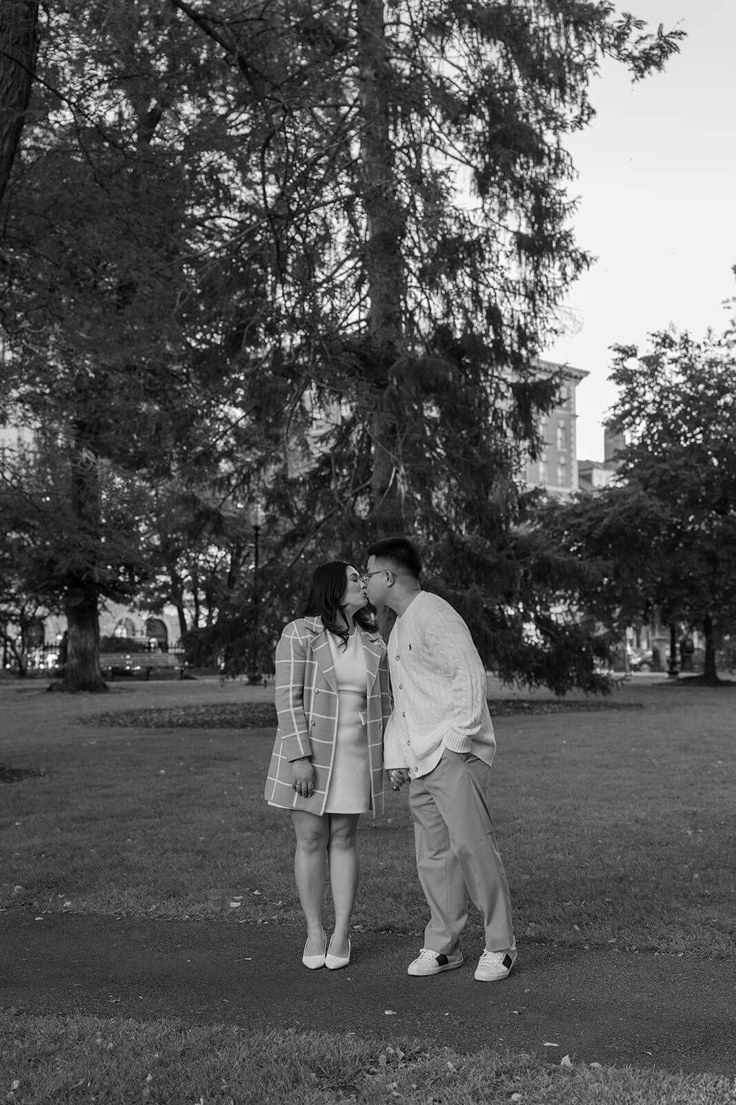 Black-and-white photograph of a couple kissing beneath tall trees in Boston Public Garden.