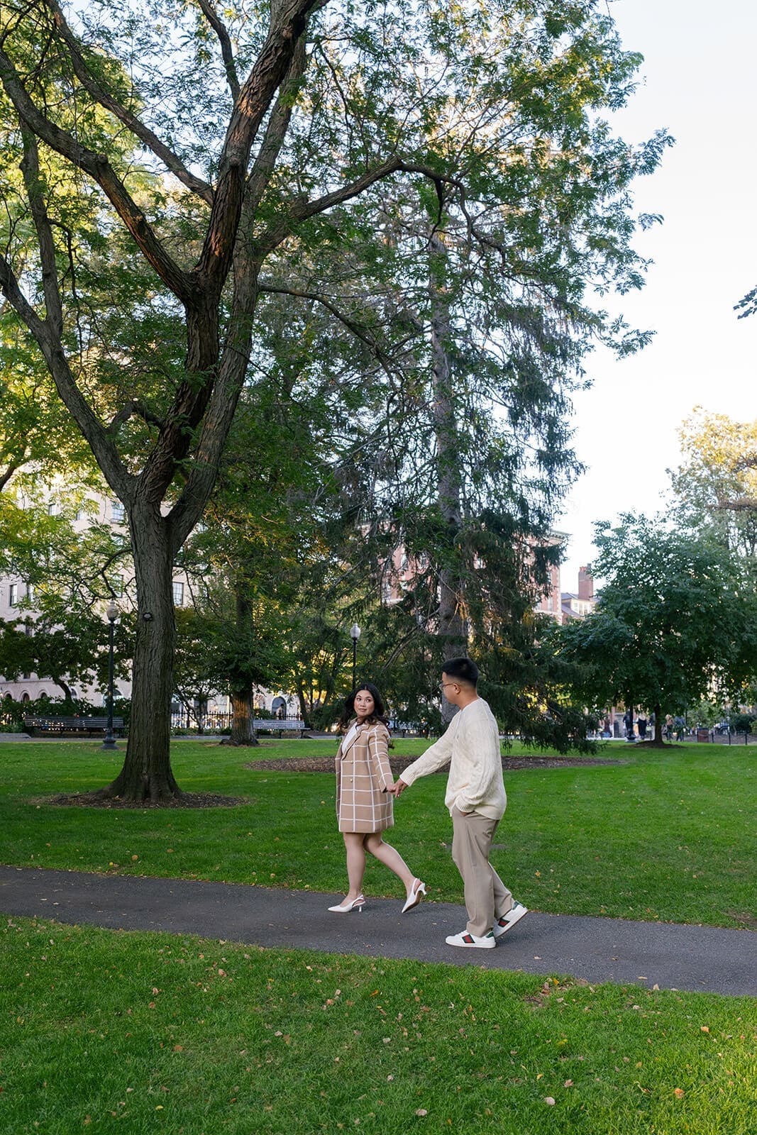 Couple walking hand in hand along a path beneath a large tree in Boston Public Garden.