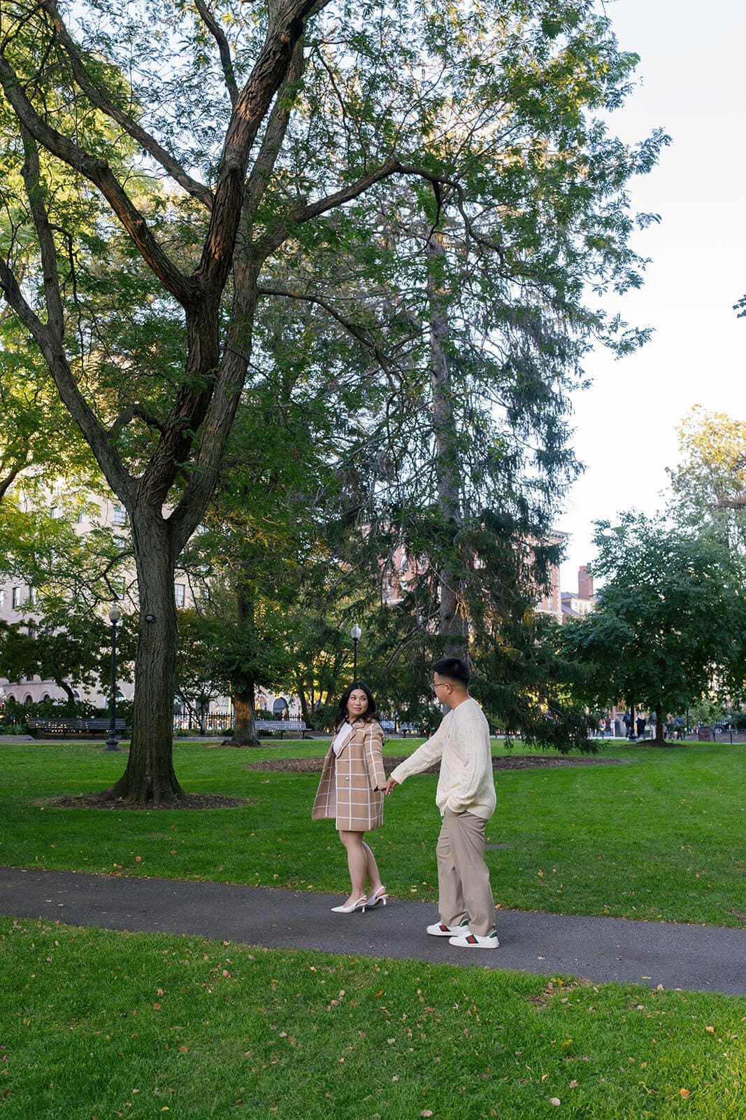 Couple reaching toward each other beneath a large tree while walking through Boston Public Garden.