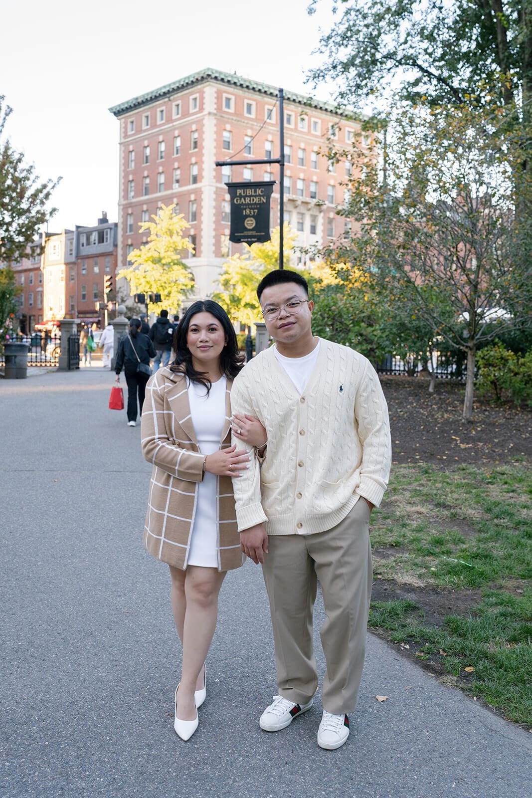Couple standing arm in arm on a path in Boston Public Garden with city buildings in the background.