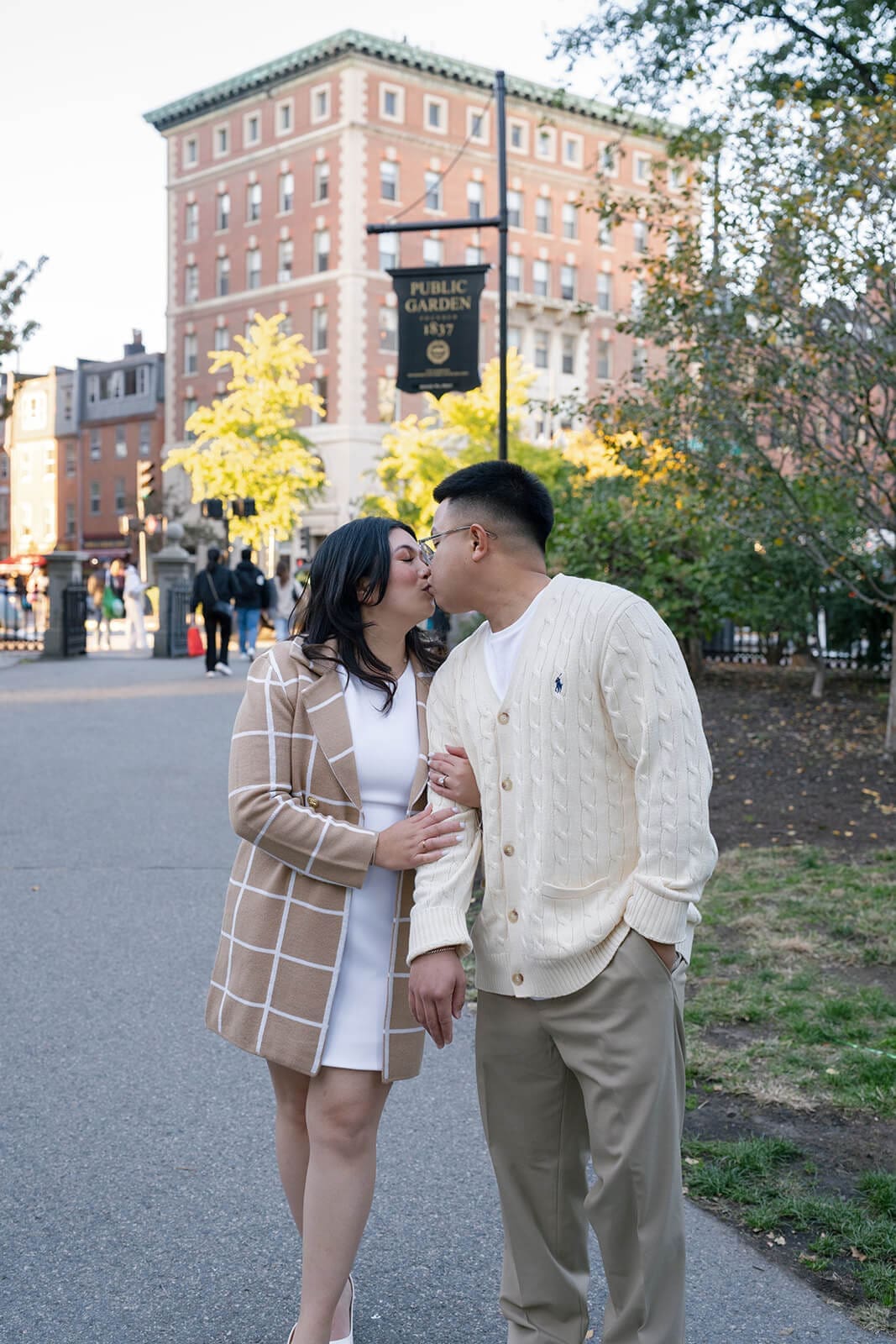 Couple kissing while walking together near the entrance to Boston Public Garden with city buildings in the background.