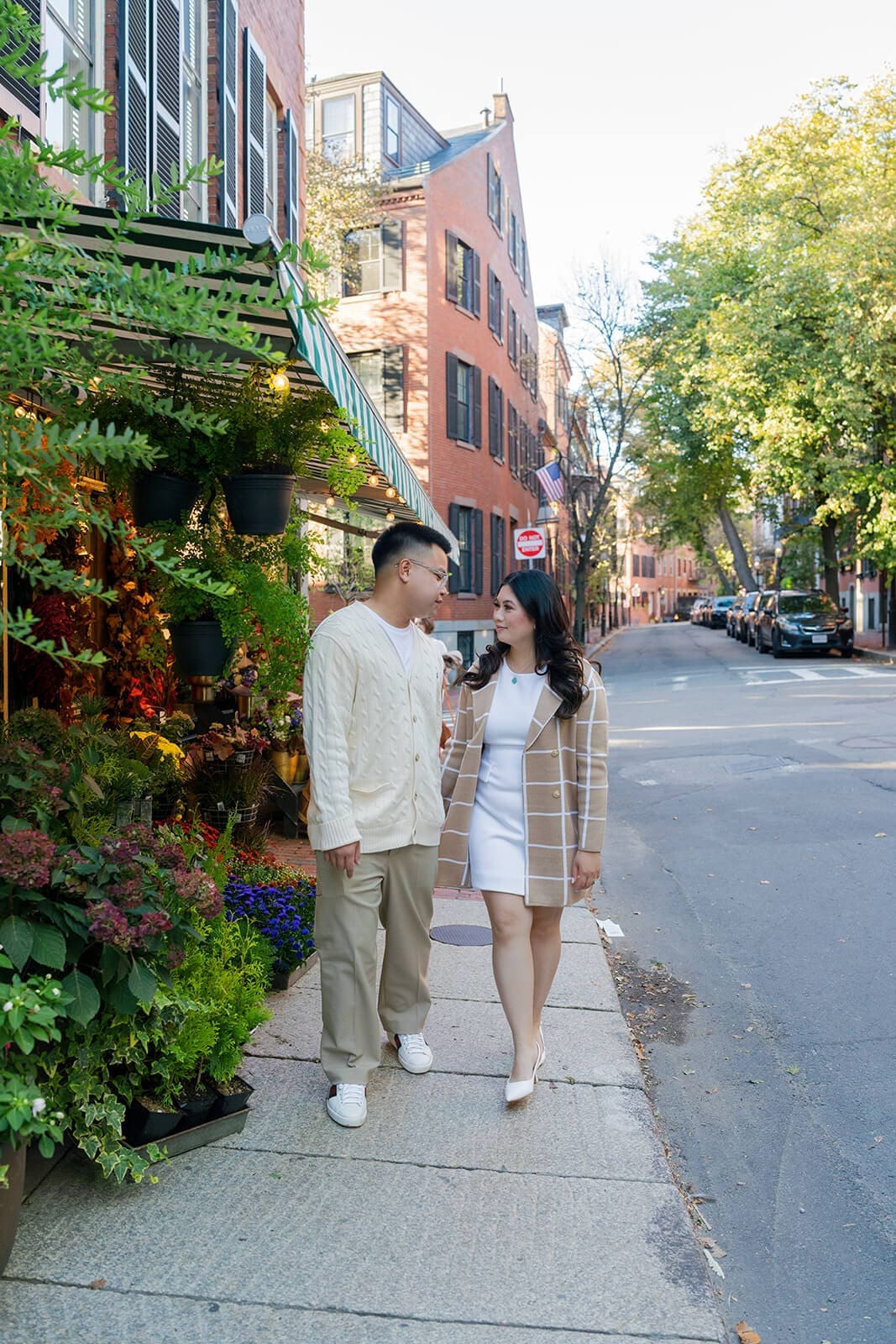 Couple walking together along the sidewalk beside a flower shop on a sunny Beacon Hill street.