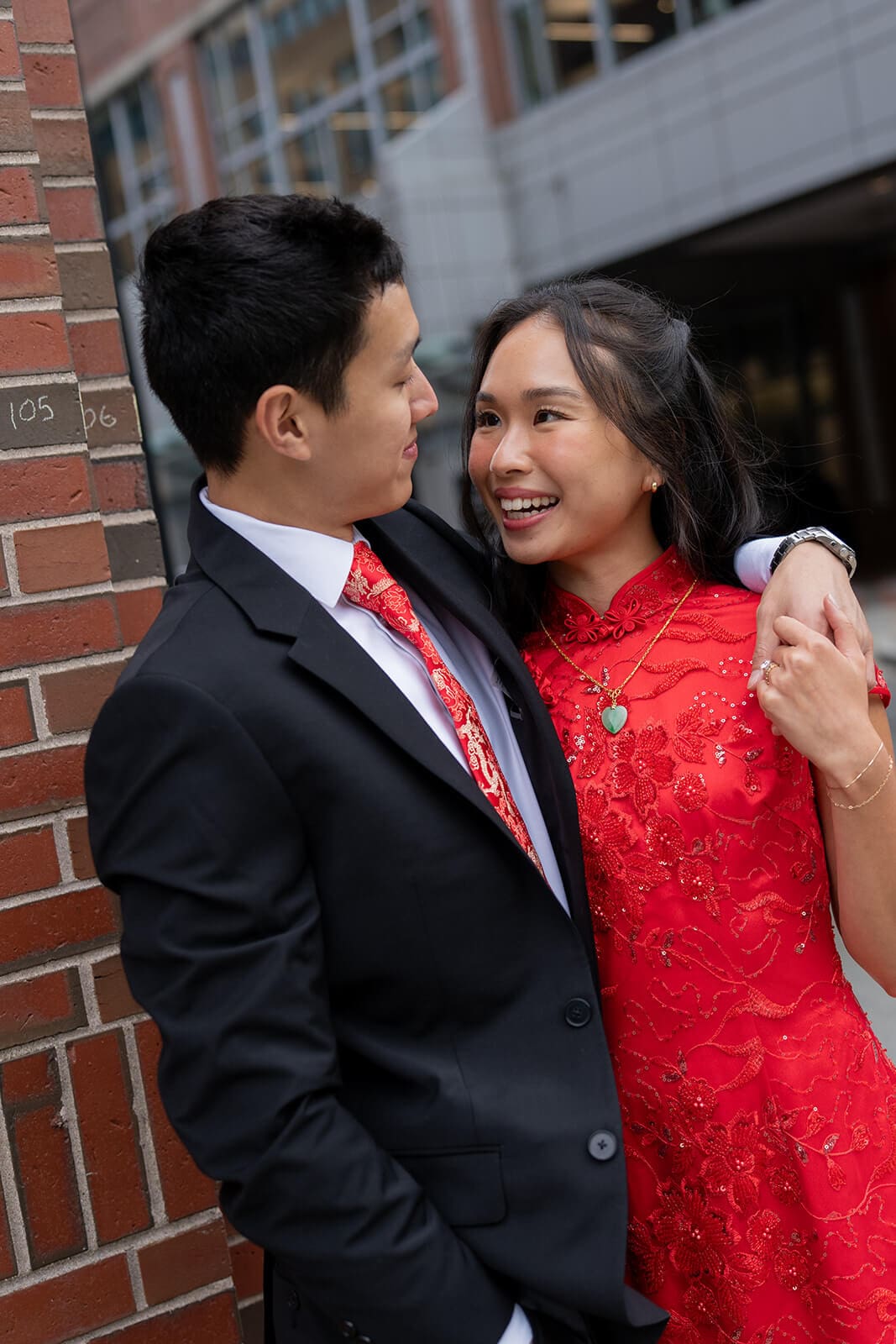 Couple smiling at each other while standing closely together beside a brick wall.