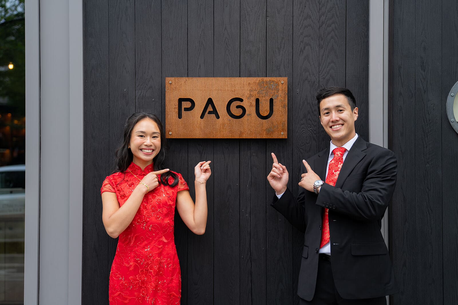Couple in red and black formal attire pointing toward the PAGU sign during their pagu restaurant wedding in Cambridge, Massachusetts.