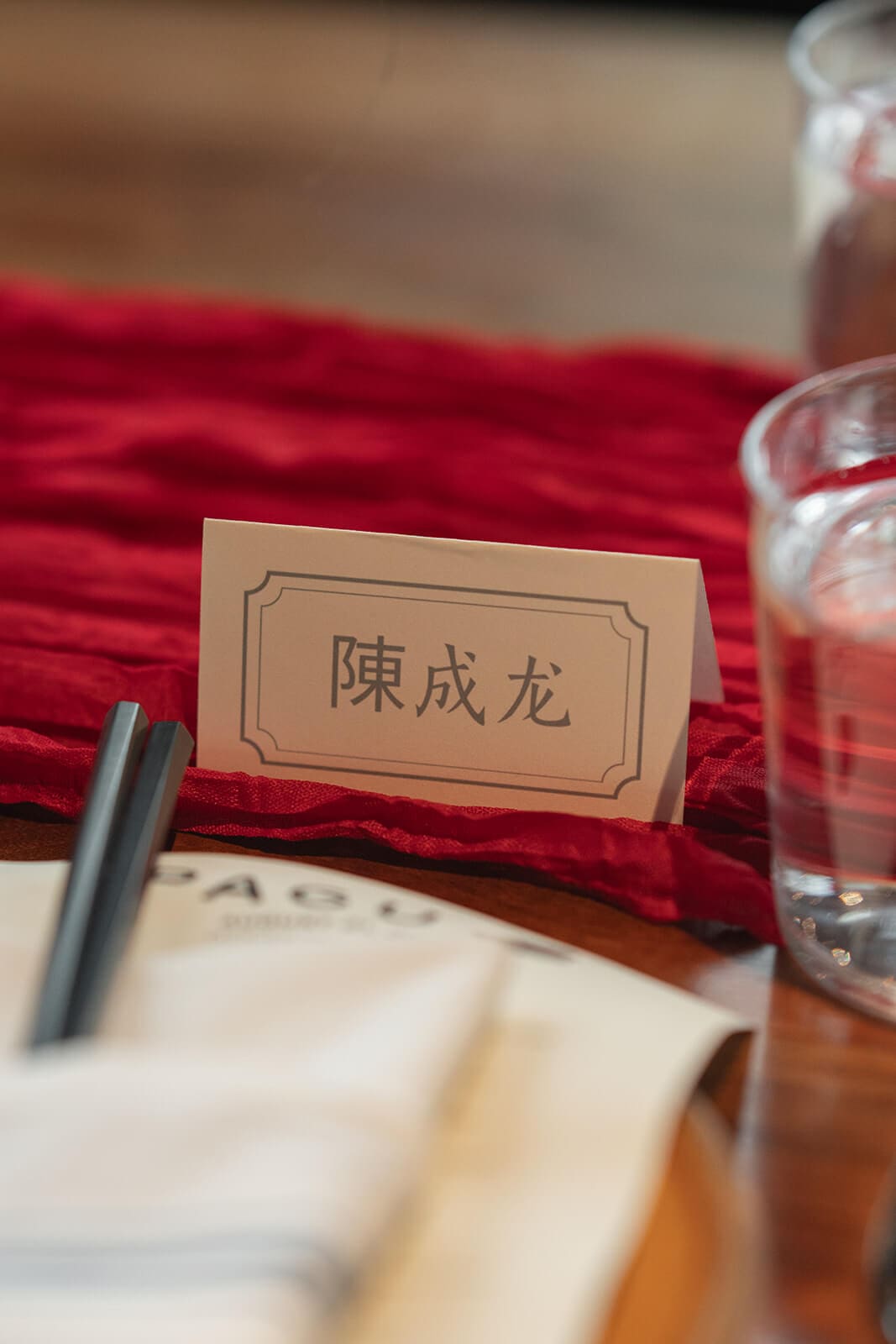 Guest place card with Chinese characters displayed on a red table runner beside chopsticks and glassware.