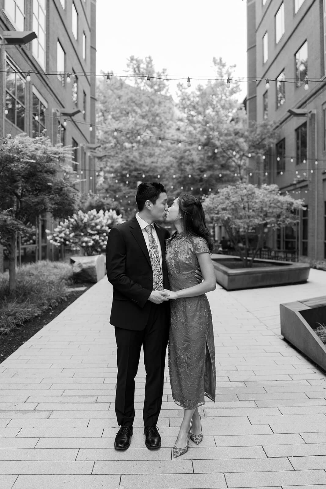 Black-and-white portrait of the couple kissing while standing together in a modern courtyard.