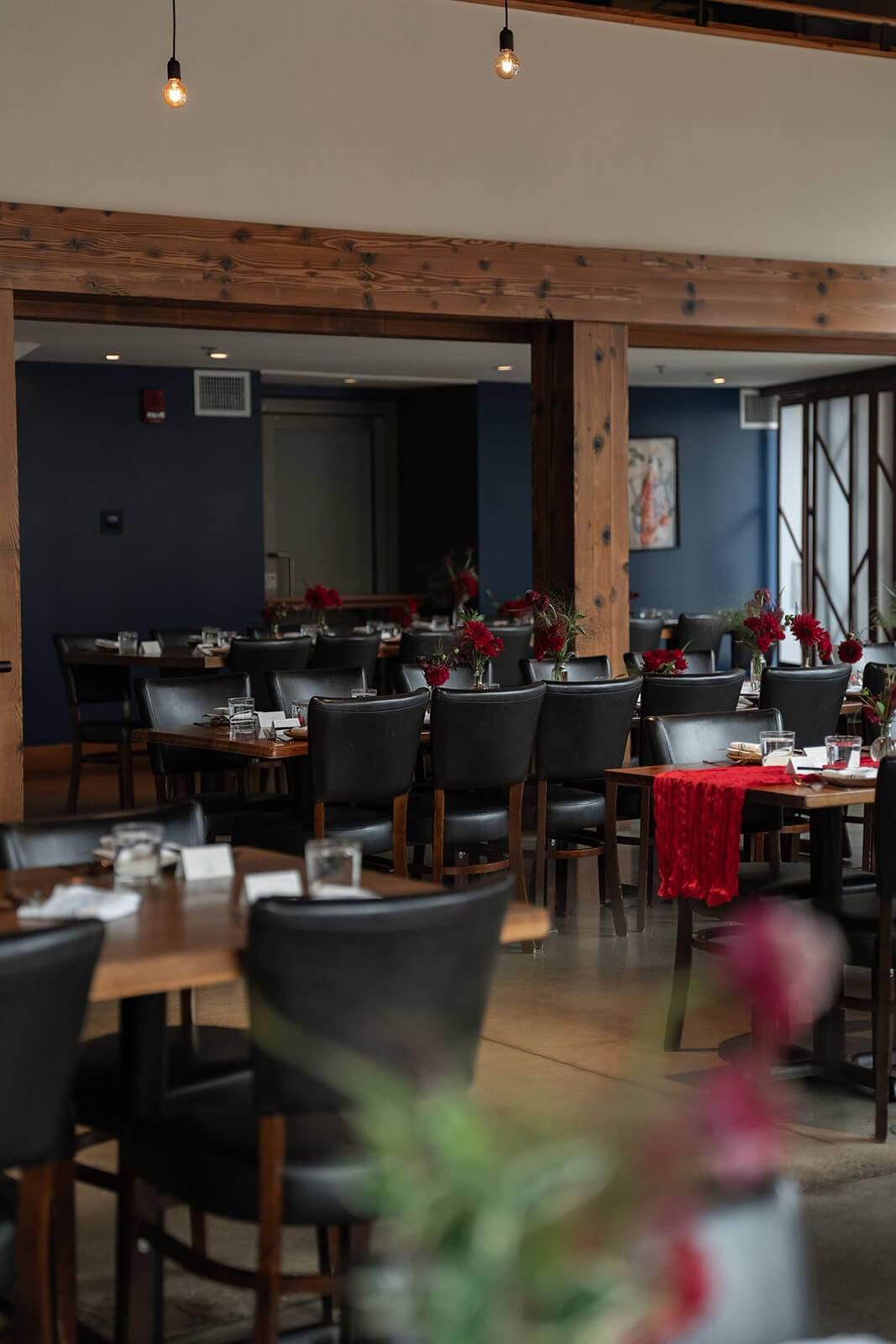 Dining room set for a pagu restaurant wedding with red floral centerpieces, dark chairs, and wooden tables.