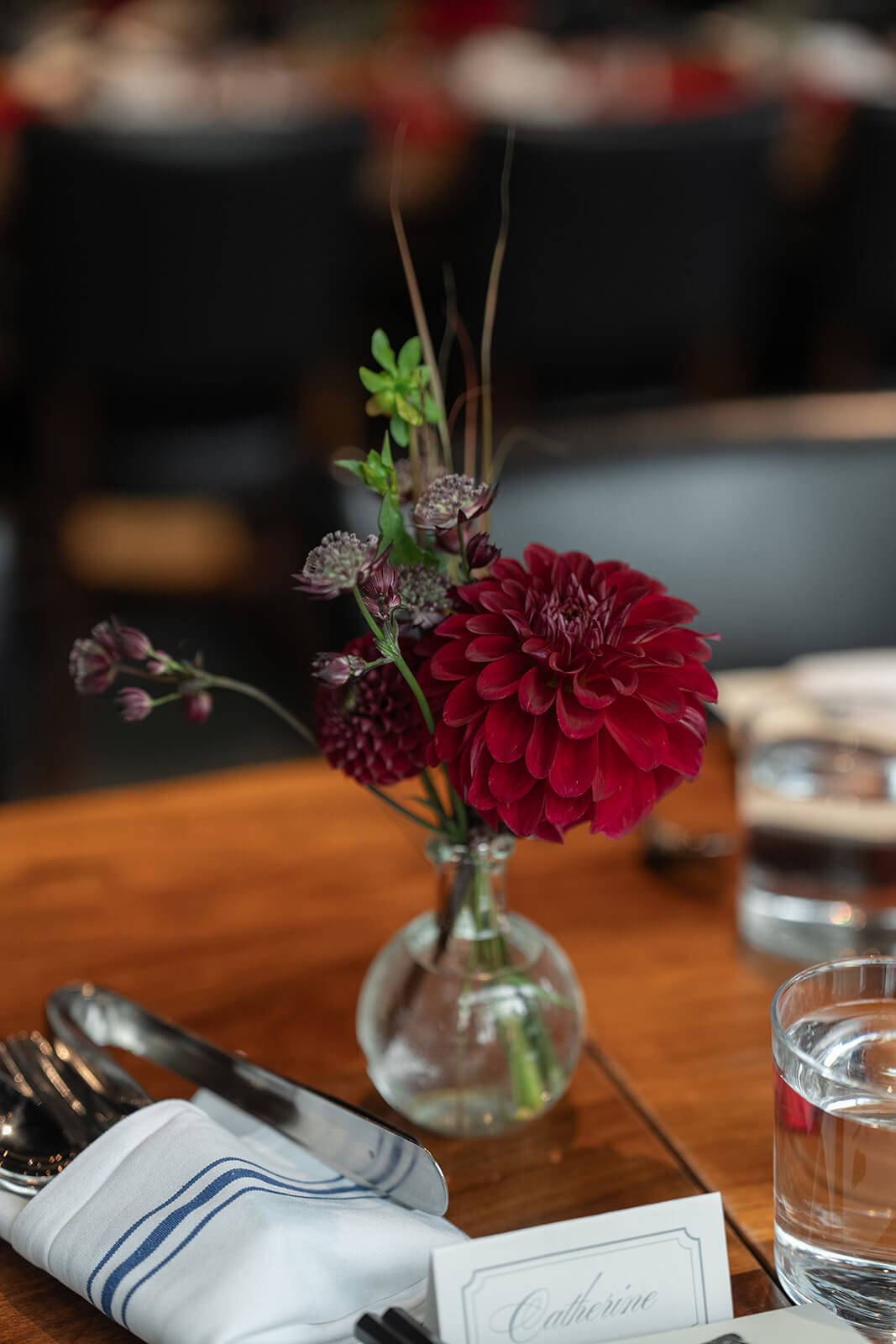 Small glass vase filled with a deep red flower and greenery beside a place setting.