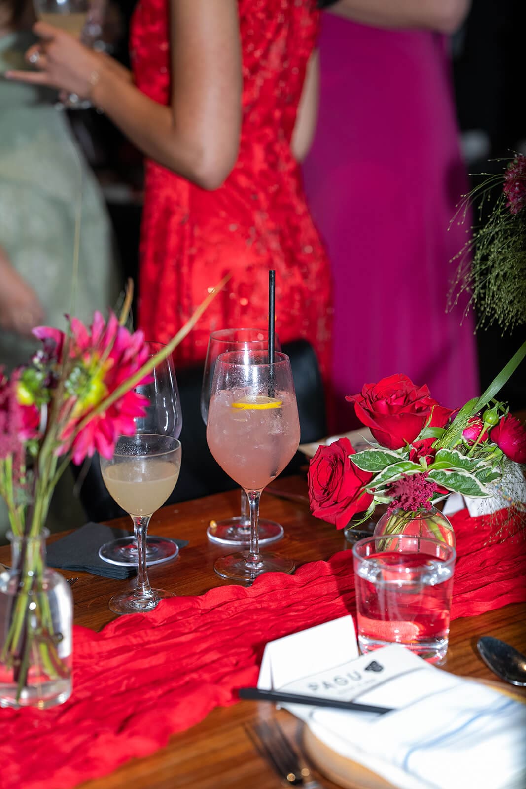 Colorful cocktails and red floral arrangements covering a reception table as guests mingle nearby.