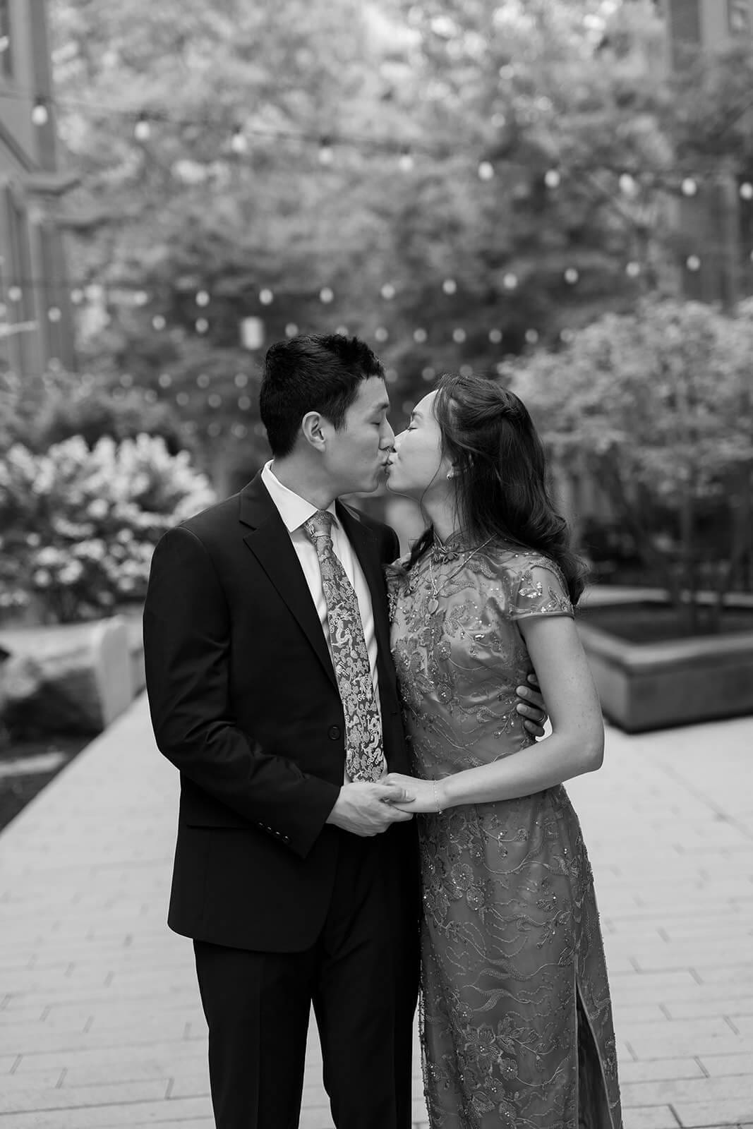 Black-and-white portrait of the couple kissing while holding hands in a landscaped courtyard.