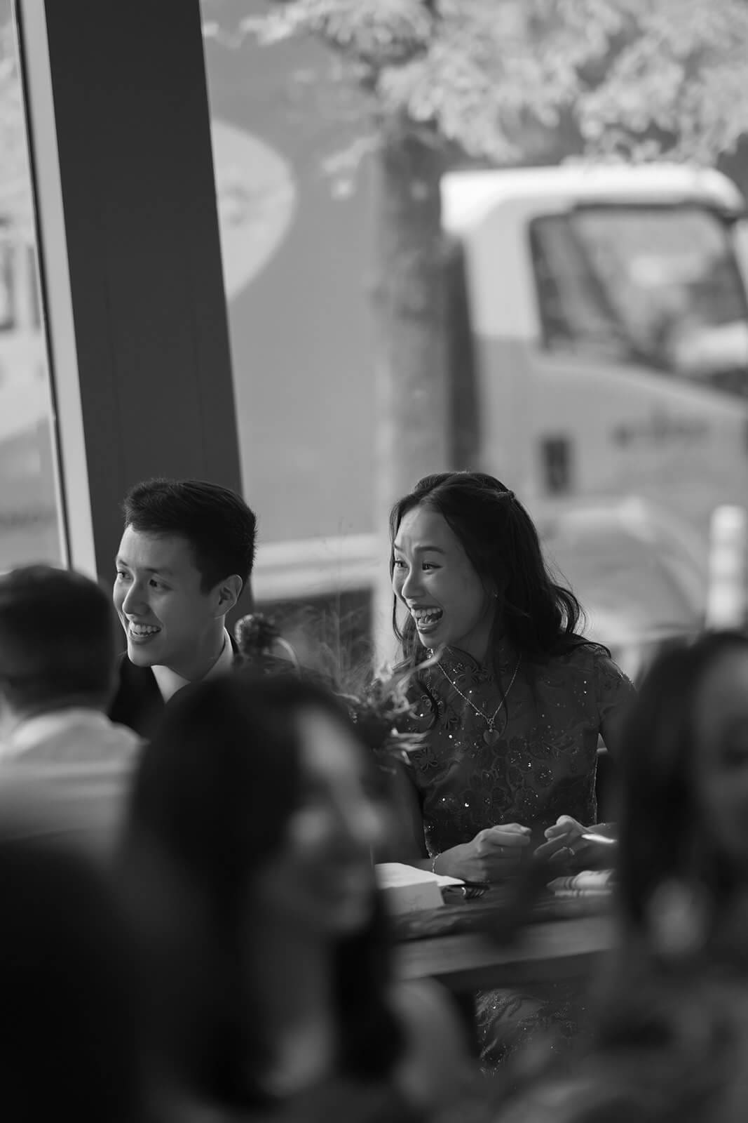Black-and-white candid of the couple laughing together at their pagu restaurant wedding dinner table.