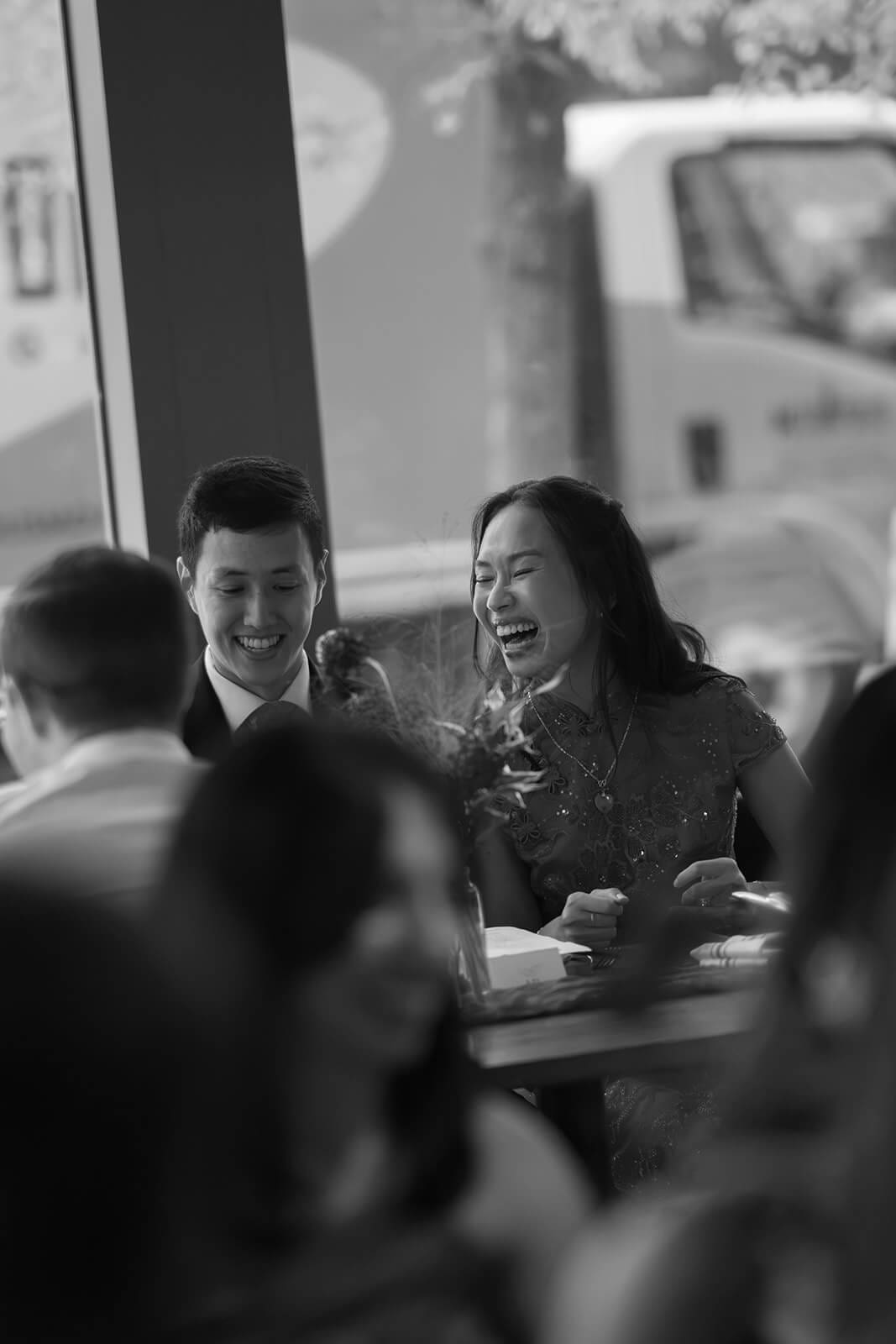 Black-and-white candid of the couple laughing together while seated among guests at their pagu restaurant wedding dinner.