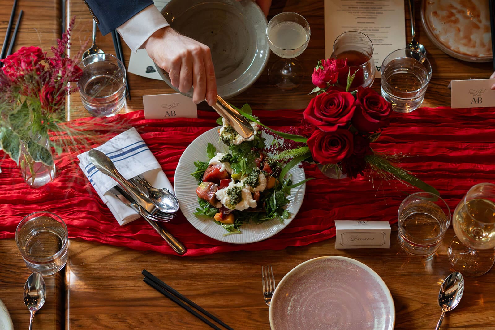 Server placing a plated salad onto a reception table surrounded by red roses, glassware, and silverware.