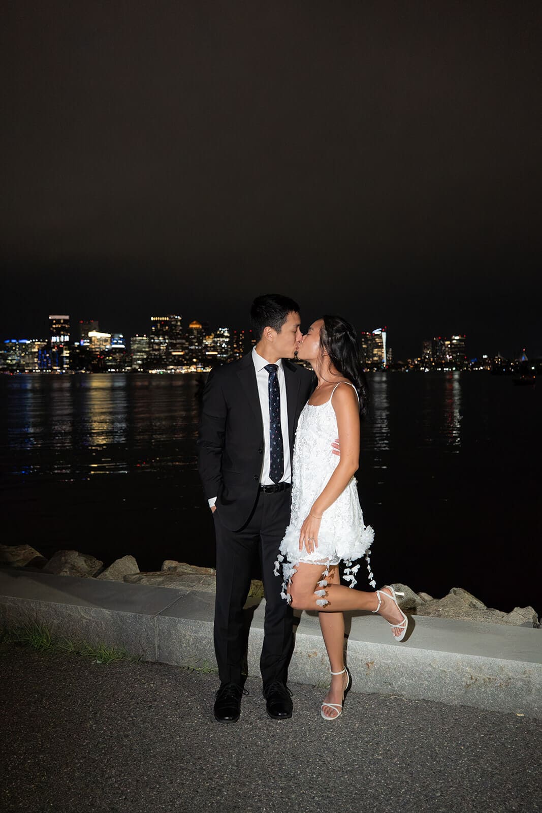 Newlyweds kissing beside the water at night with the illuminated Boston skyline behind them.