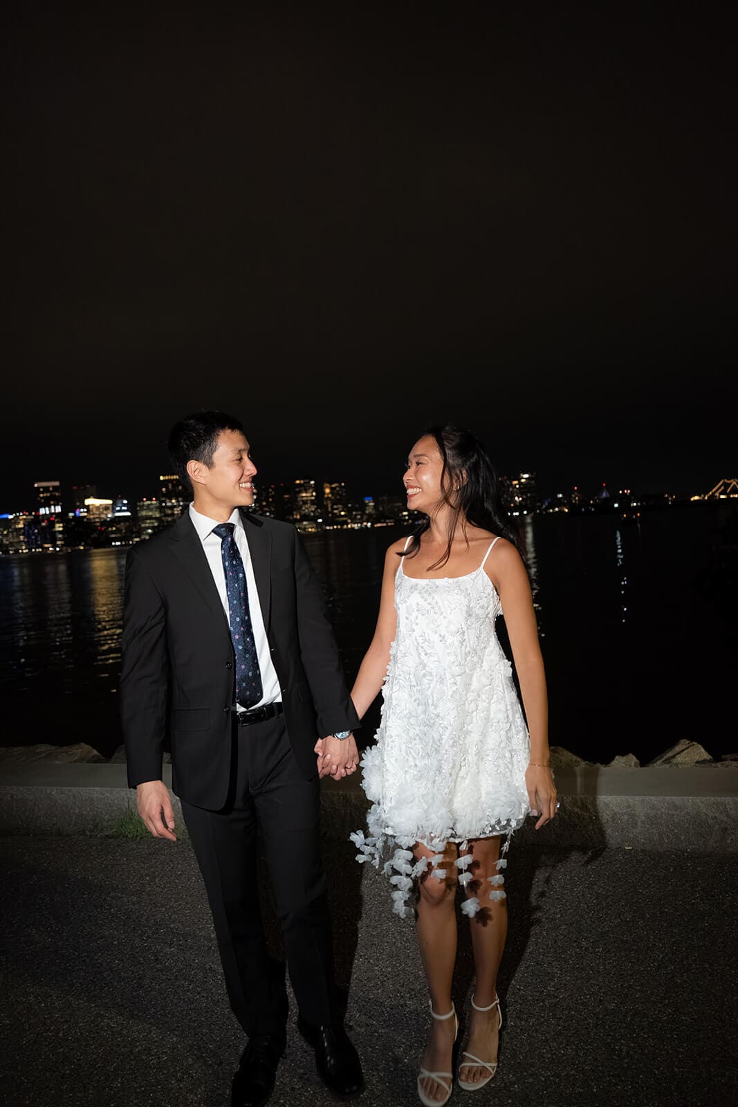 Newlyweds holding hands beside the waterfront at night with the city skyline glowing across the water.