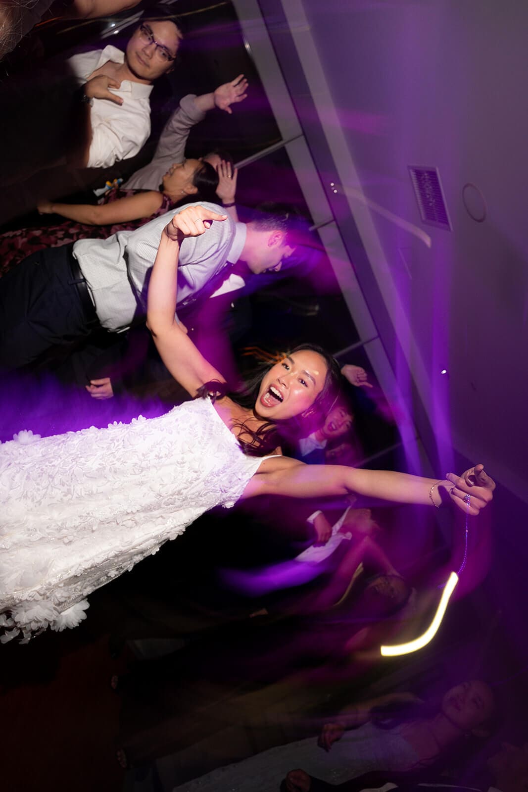 Woman in a white reception dress dancing energetically beneath purple party lighting.