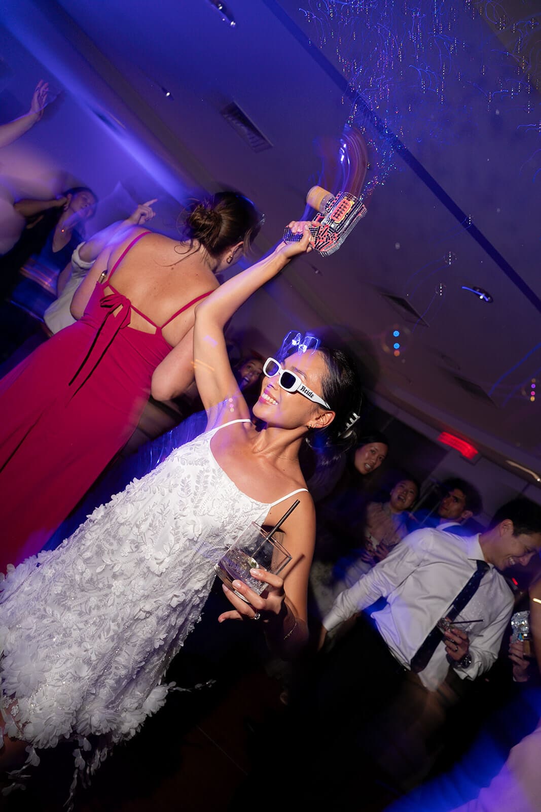 Woman in a short white textured reception dress dancing while holding a colorful toy bubble gun.