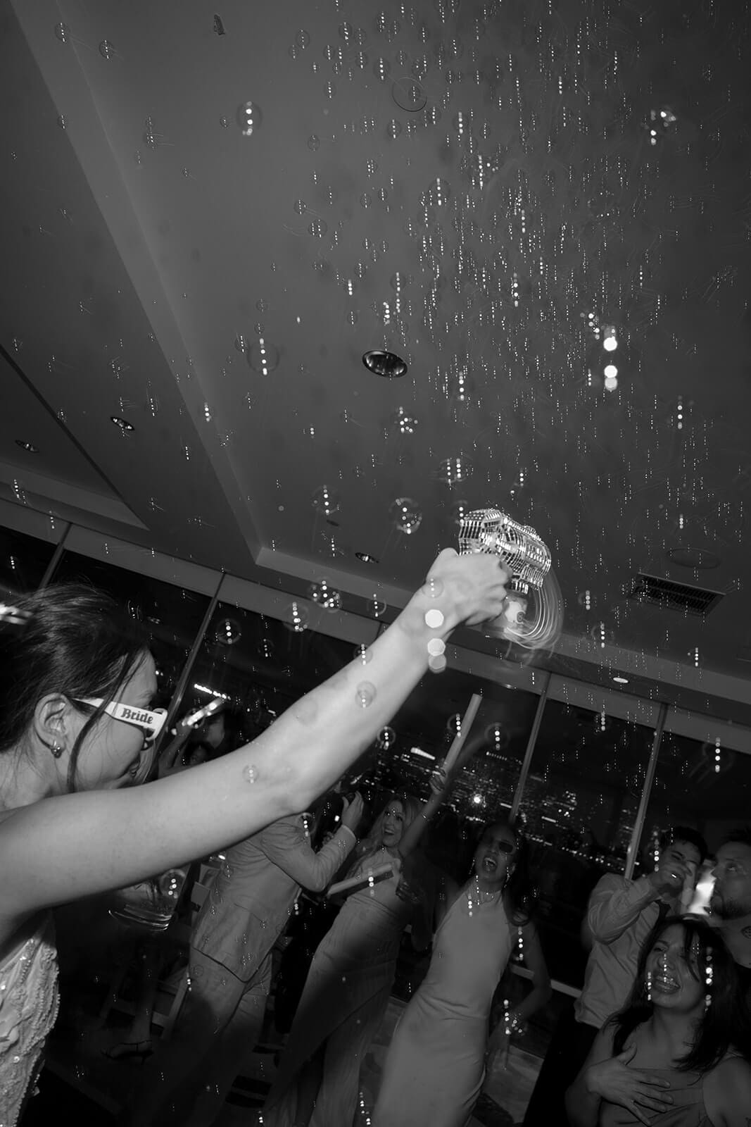Black-and-white action shot of wedding guests dancing beneath bubbles and dramatic reception lighting.