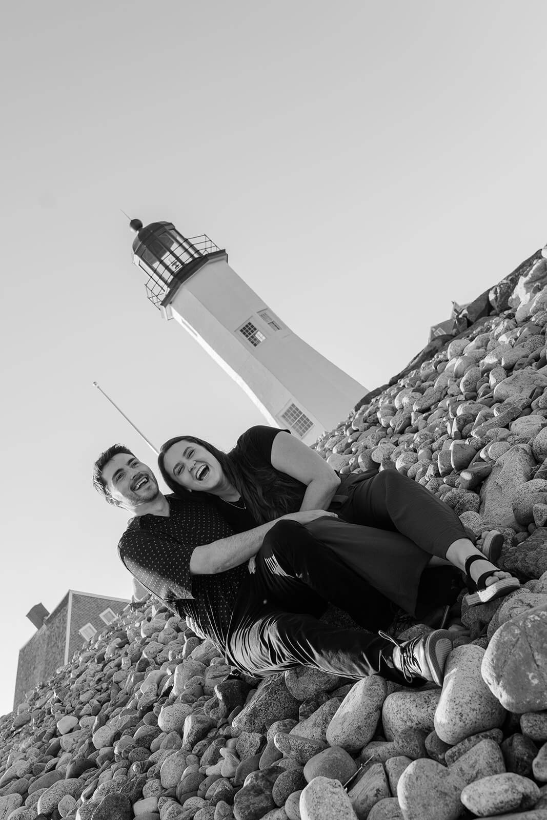 Black-and-white photo of an engaged couple laughing together on the rocky shoreline with Scituate Lighthouse behind them