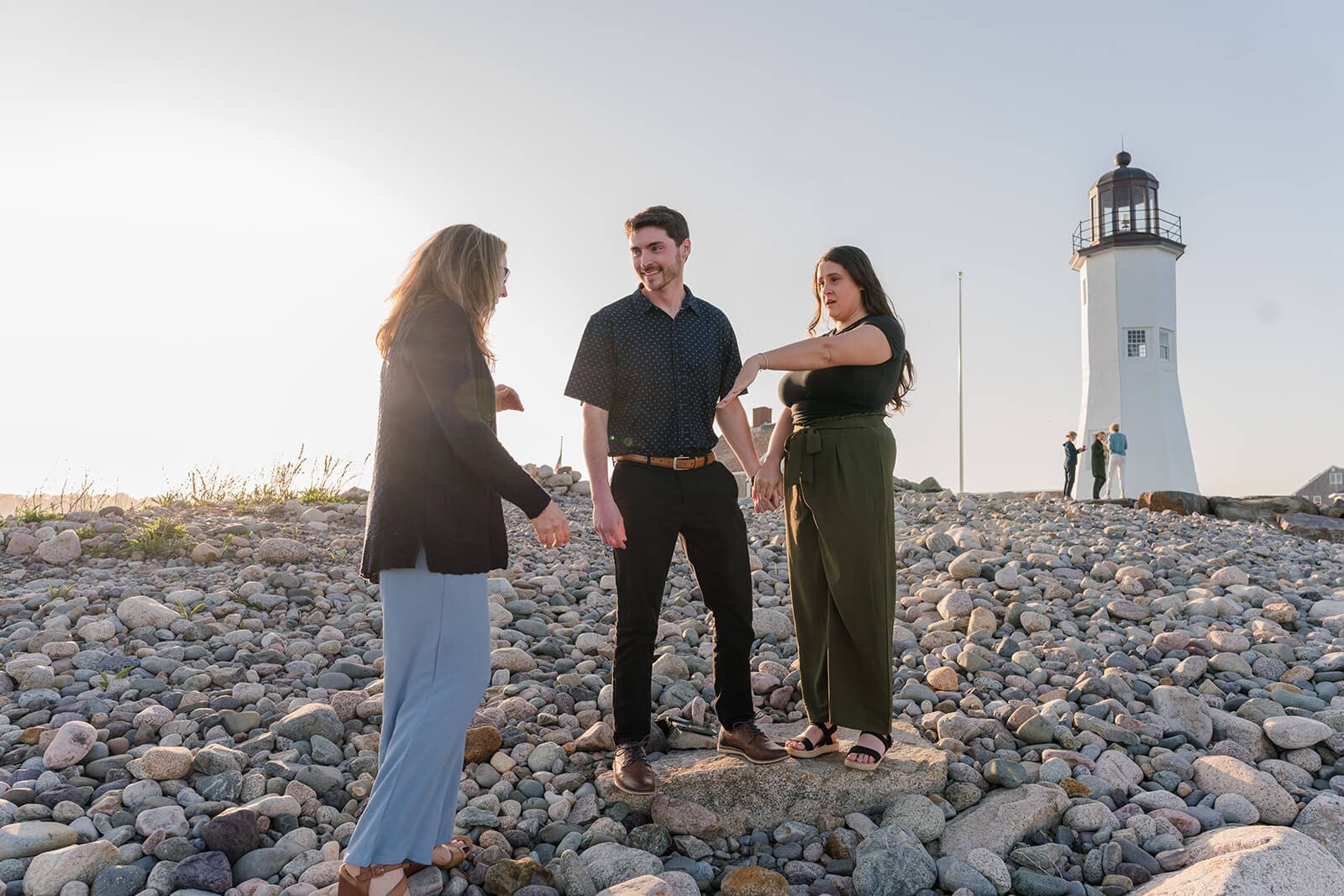 A couple talking with a family member on the rocky shoreline near Scituate Lighthouse showing them an engagement ring