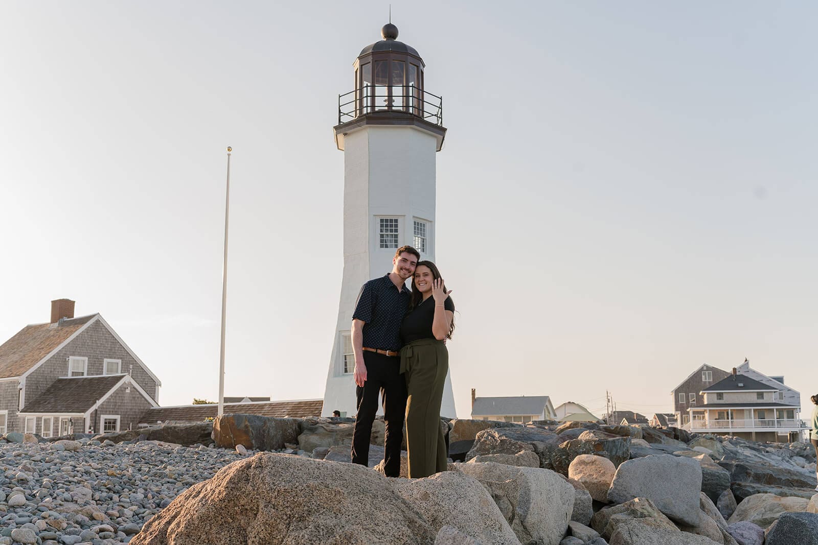 Engaged couple standing together on large coastal rocks in front of Scituate Lighthouse