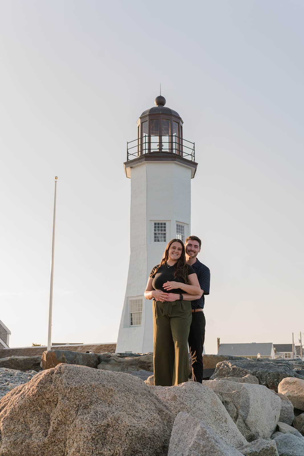 Couple embracing on a large rock with Scituate Lighthouse behind them